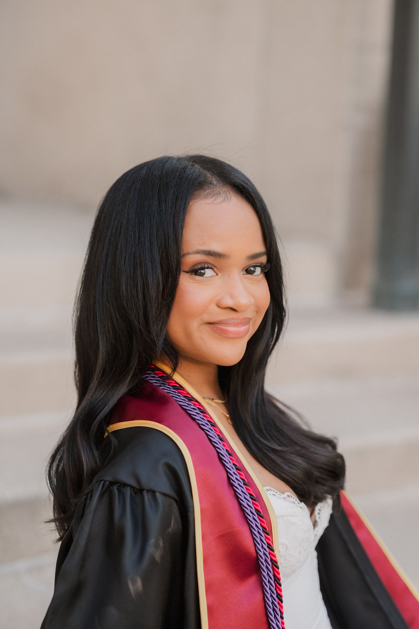 Young woman smiling in black and red graduation gown with honor cords, posing outdoors for graduation photo.