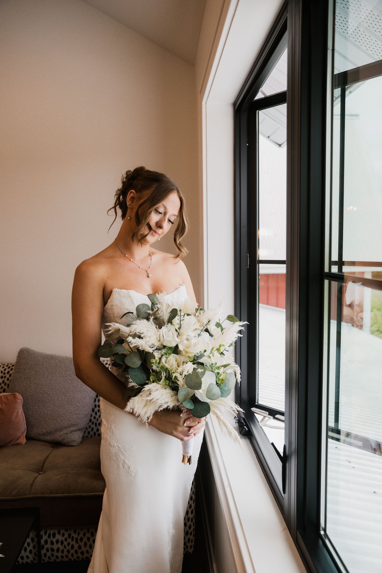 Bride in strapless white gown holds bouquet of white roses and eucalyptus by a sunlit window.