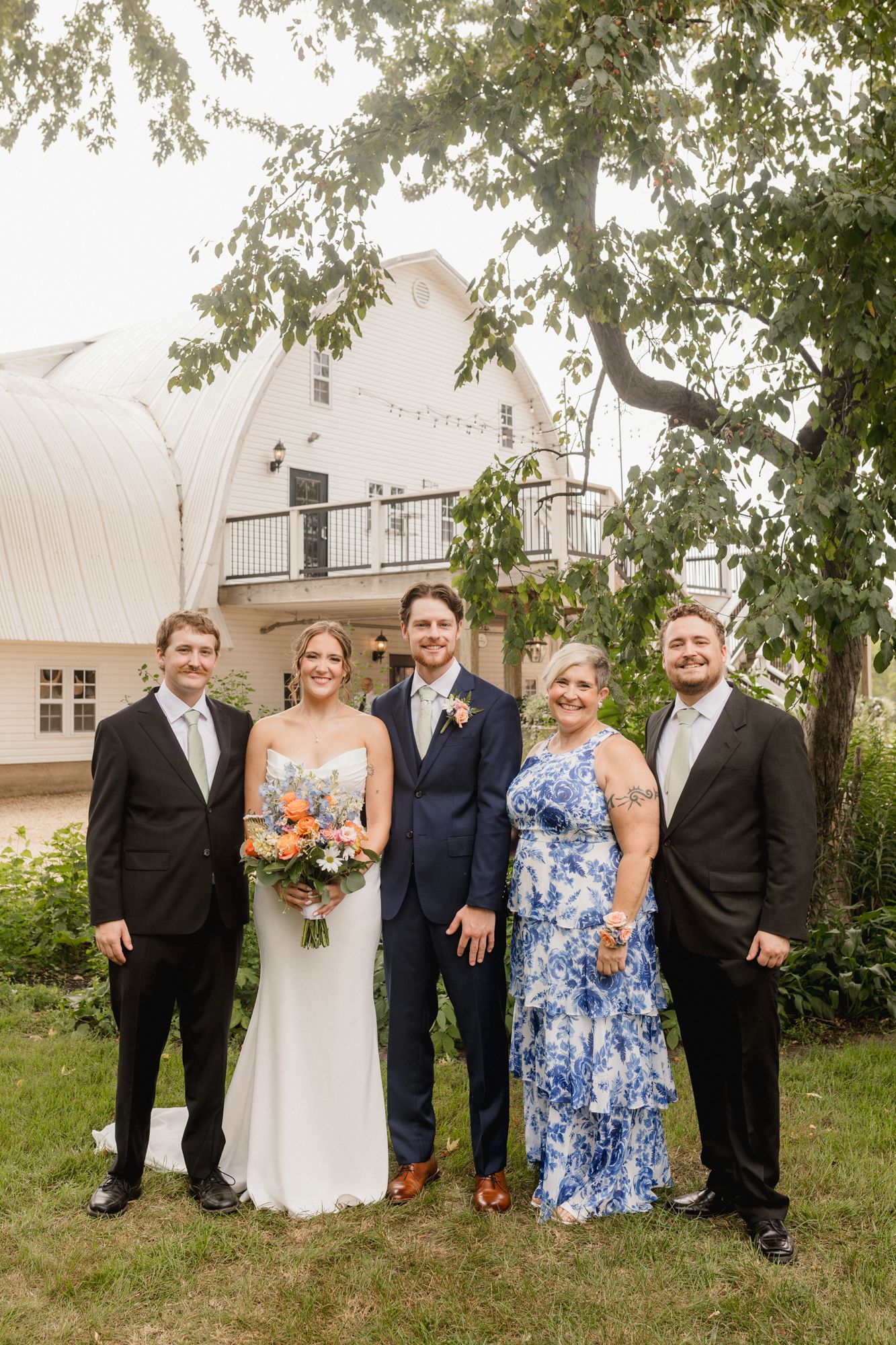 Wedding party of five poses outdoors near white barn venue, bride holding colorful bouquet with groom in navy suit.