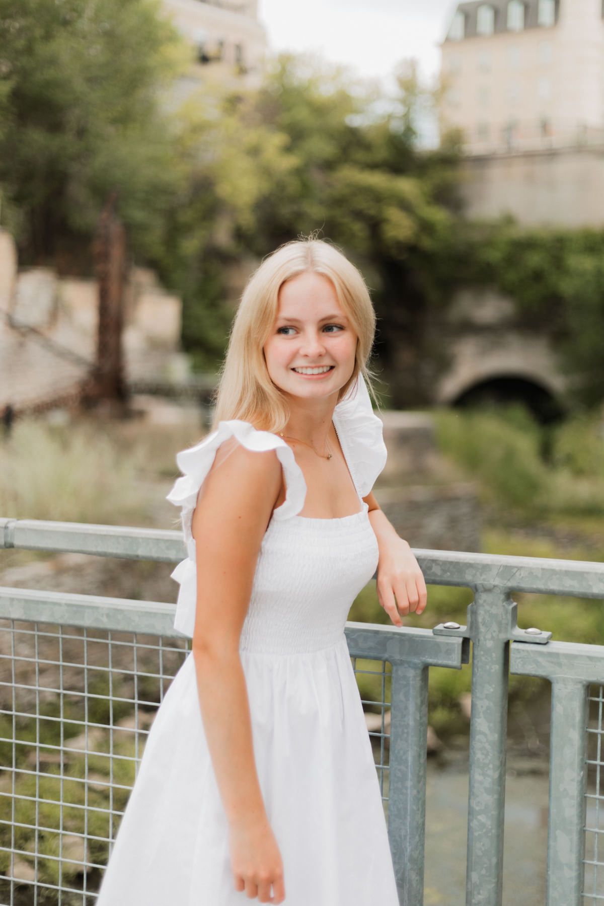 Smiling blonde young woman in white ruffled sundress standing on bridge with green trees in background.