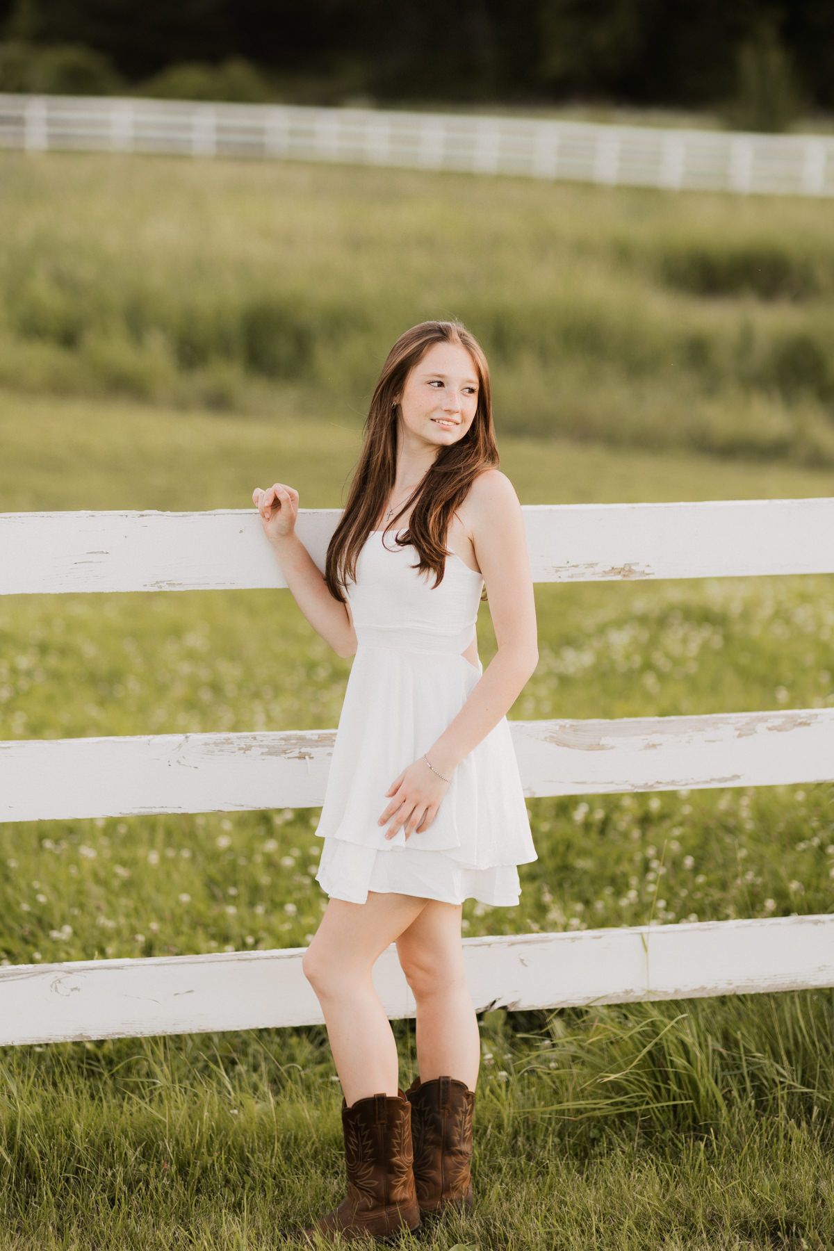 Young woman in white dress and cowboy boots leaning against a white wooden fence in a green field.