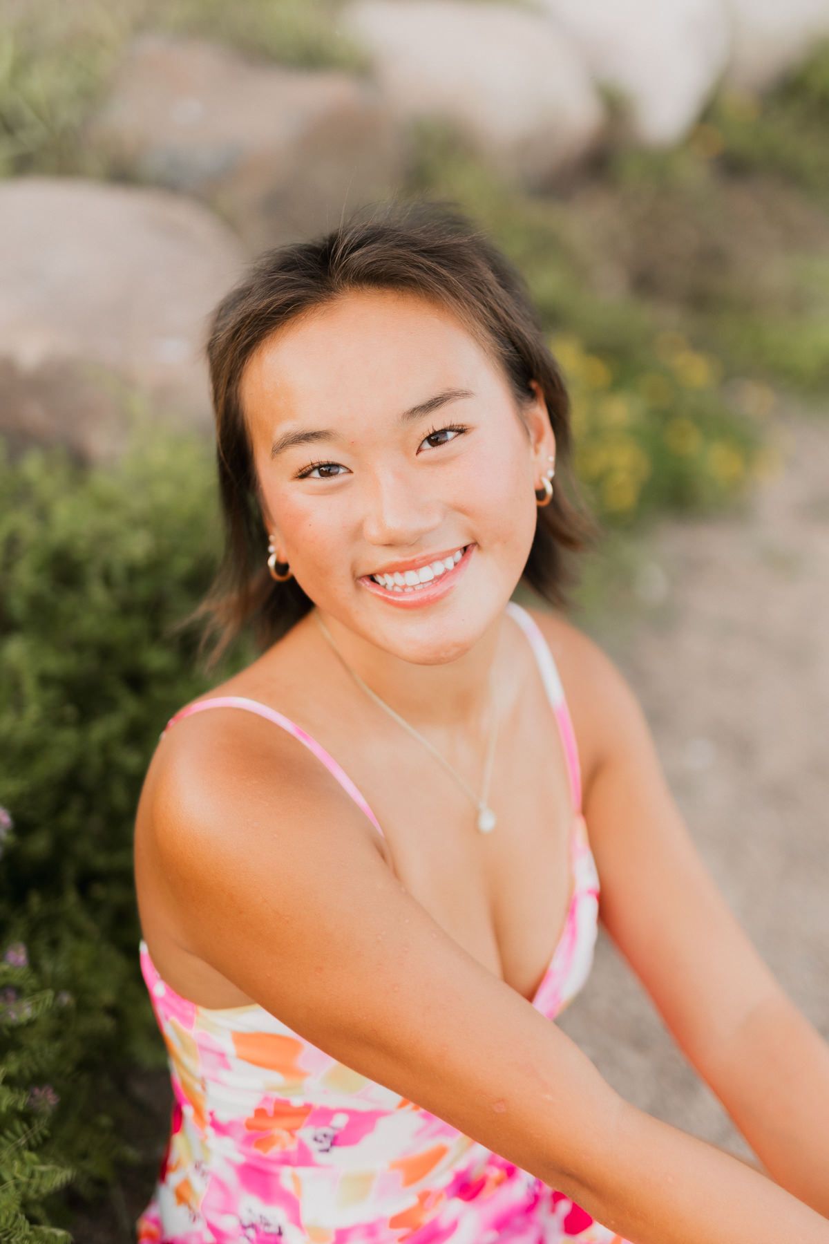 Smiling young woman in floral pink dress with necklace sitting outdoors near rocks.