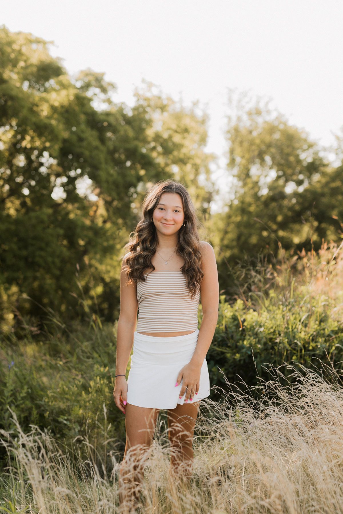 Young woman in beige crop top and white skirt smiling in golden hour sunlit meadow with tall grass.