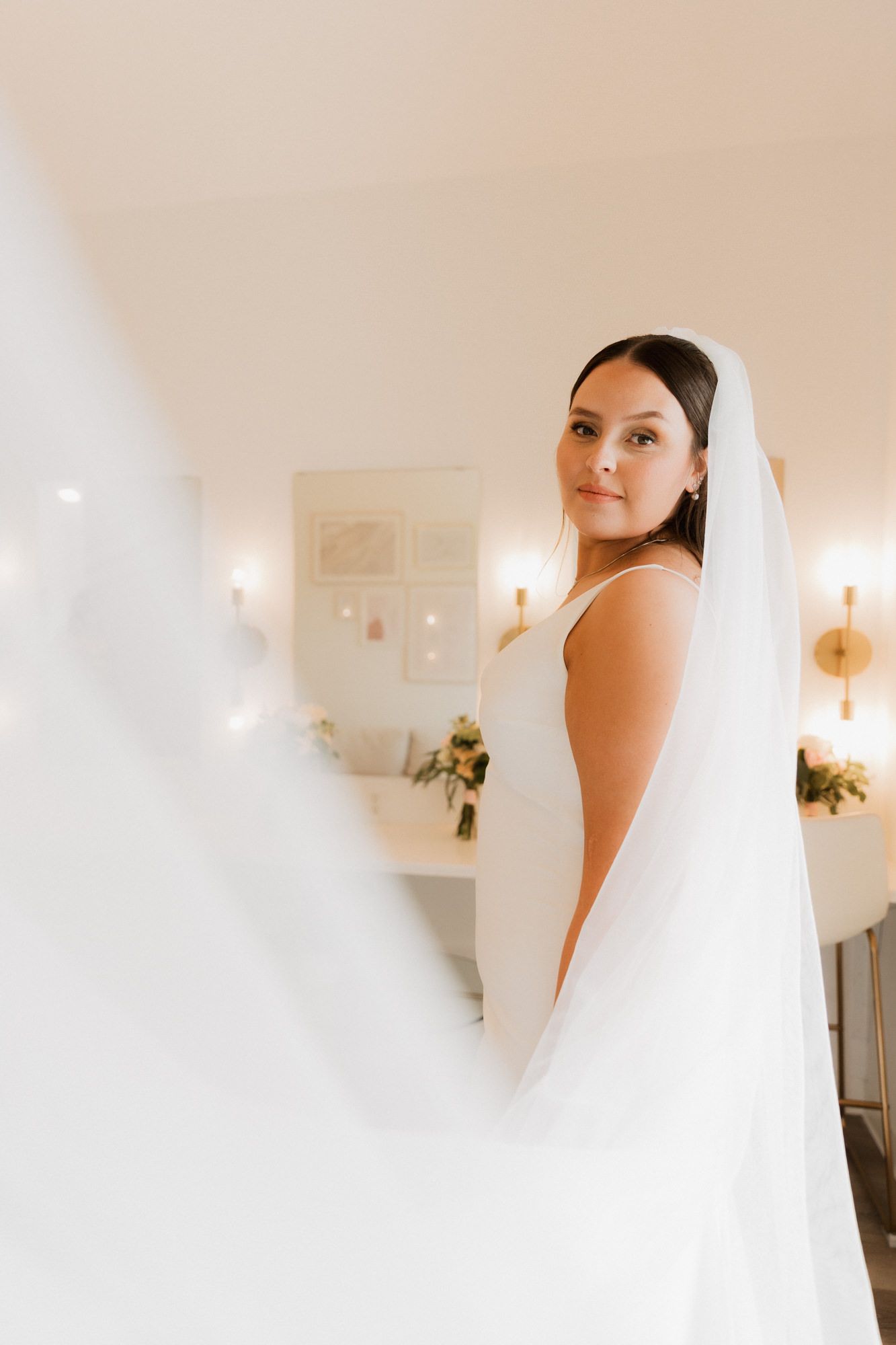 Elegant bride in a white sleeveless gown and veil gazes over her shoulder in a softly lit bridal suite.