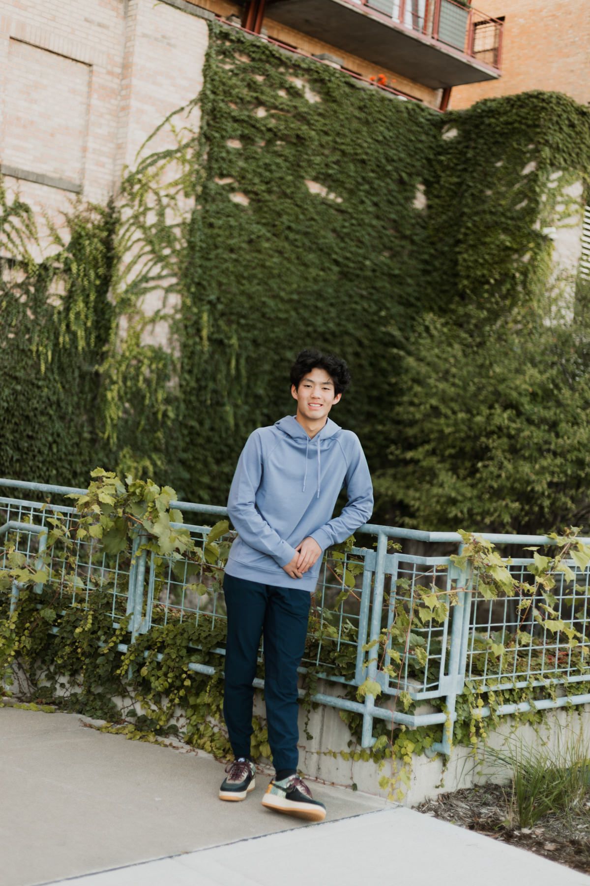 A smiling young man in a light blue hoodie and dark pants stands on a walkway surrounded by lush ivy-covered walls.