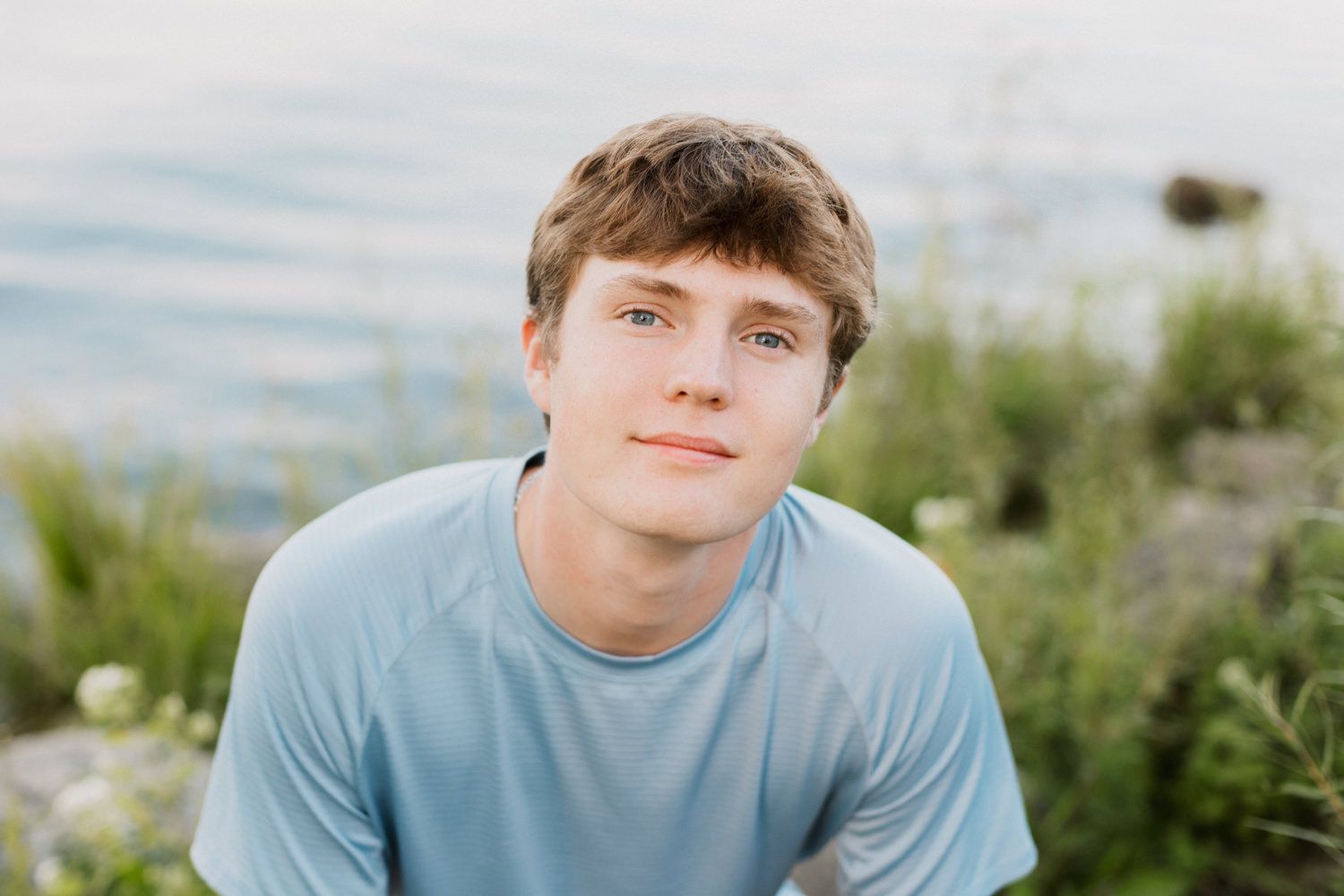 Person wearing blue shirt poses along rocky waterfront with natural scenery in background.