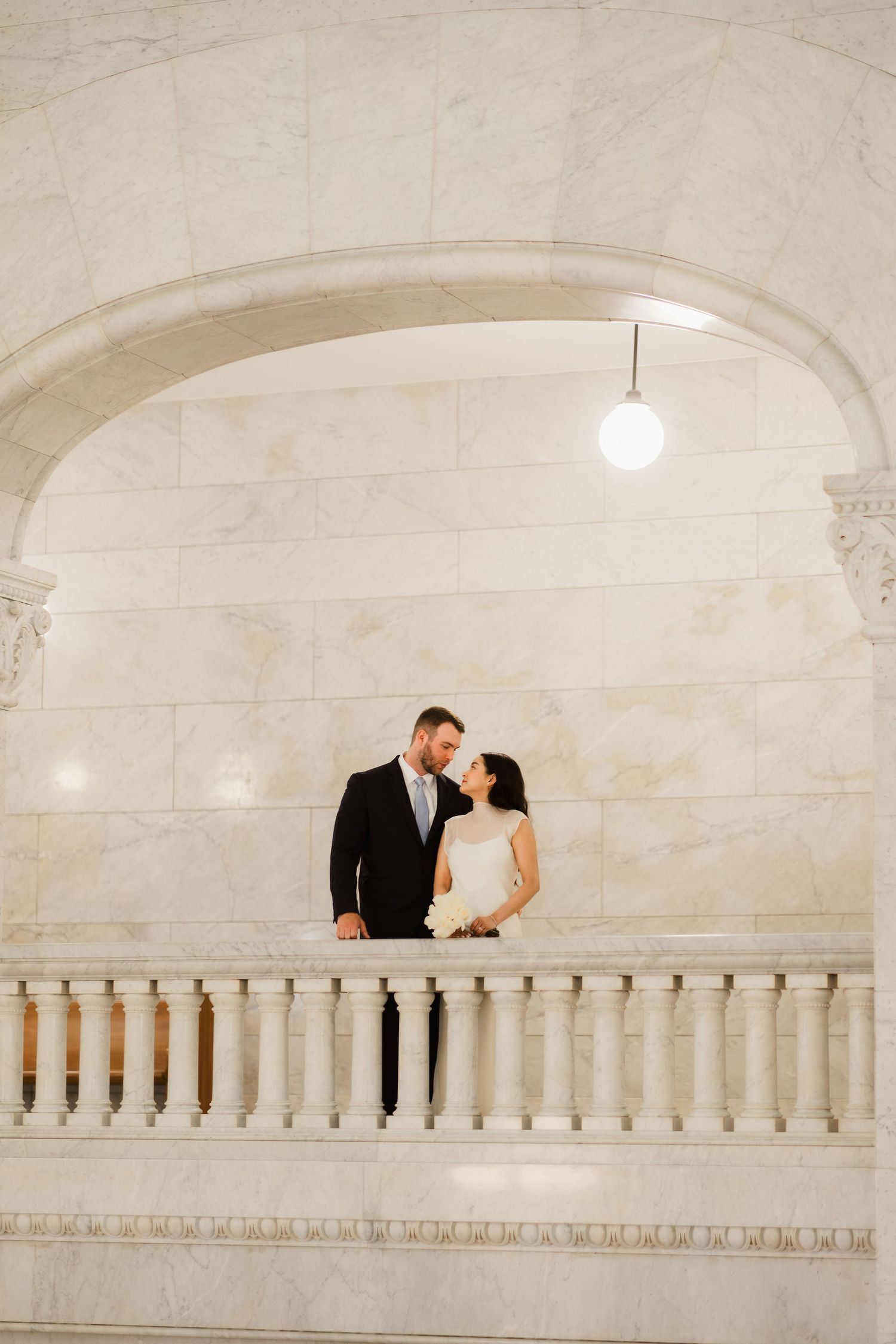Bride and groom share a romantic moment on a marble balcony under an arched ceiling in an elegant venue.
