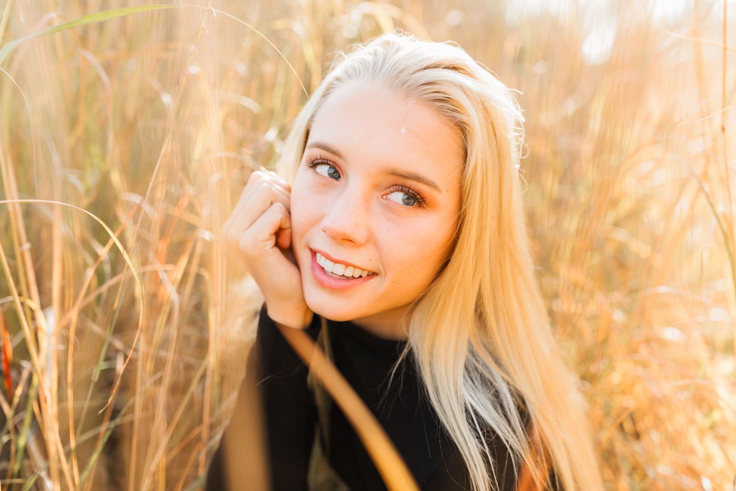 Smiling blonde teenage girl posing outdoors in golden tall grass during warm sunset light.