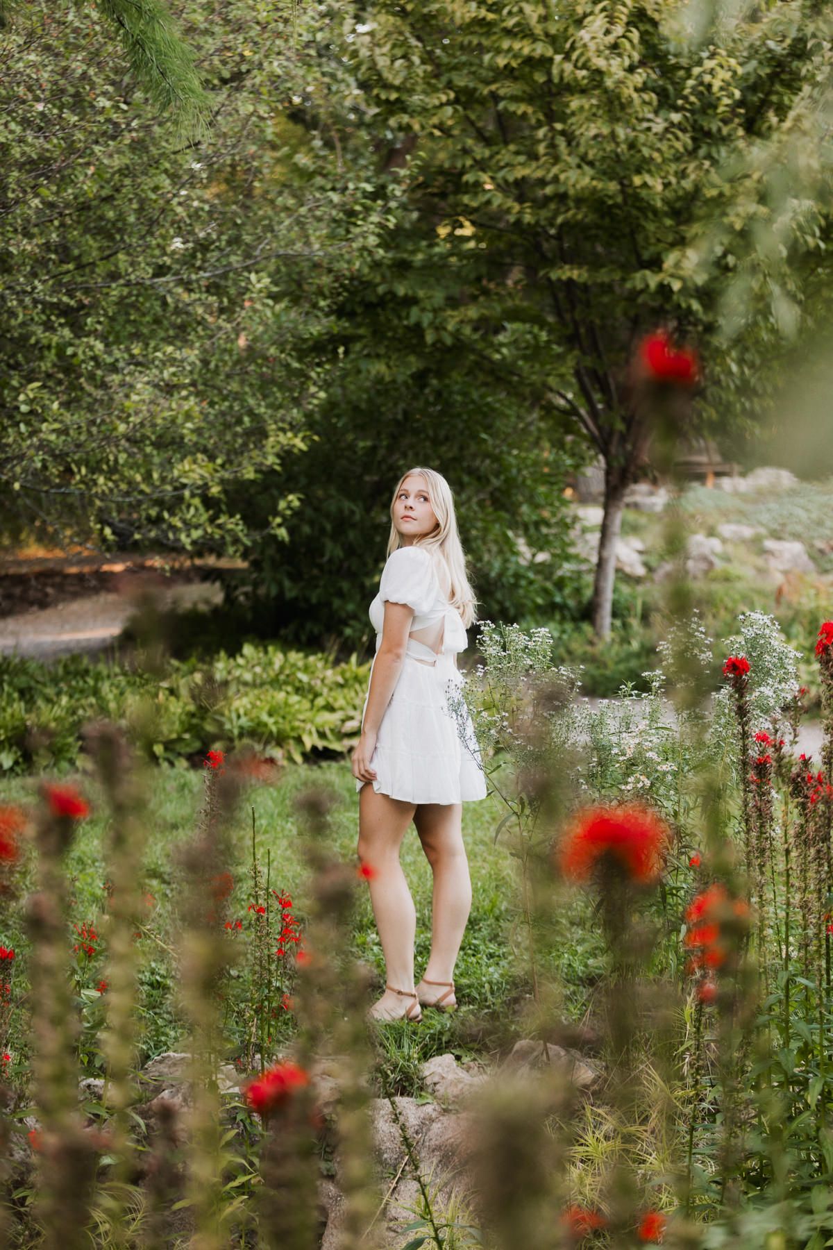 Young blonde woman in white dress standing among red flowers in a lush green garden, looking back over shoulder.