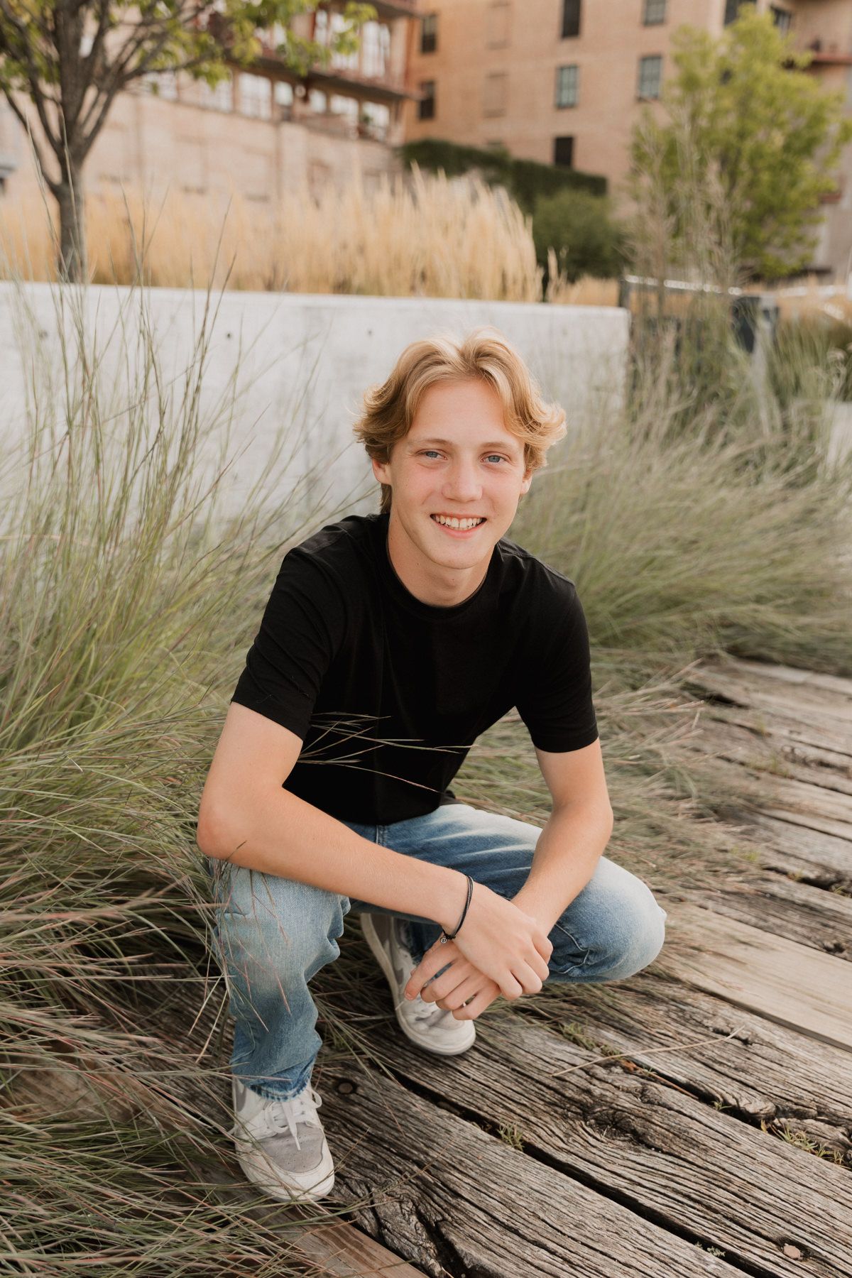 Smiling teenage boy in black t-shirt and jeans crouching on weathered wood planks surrounded by ornamental grasses.