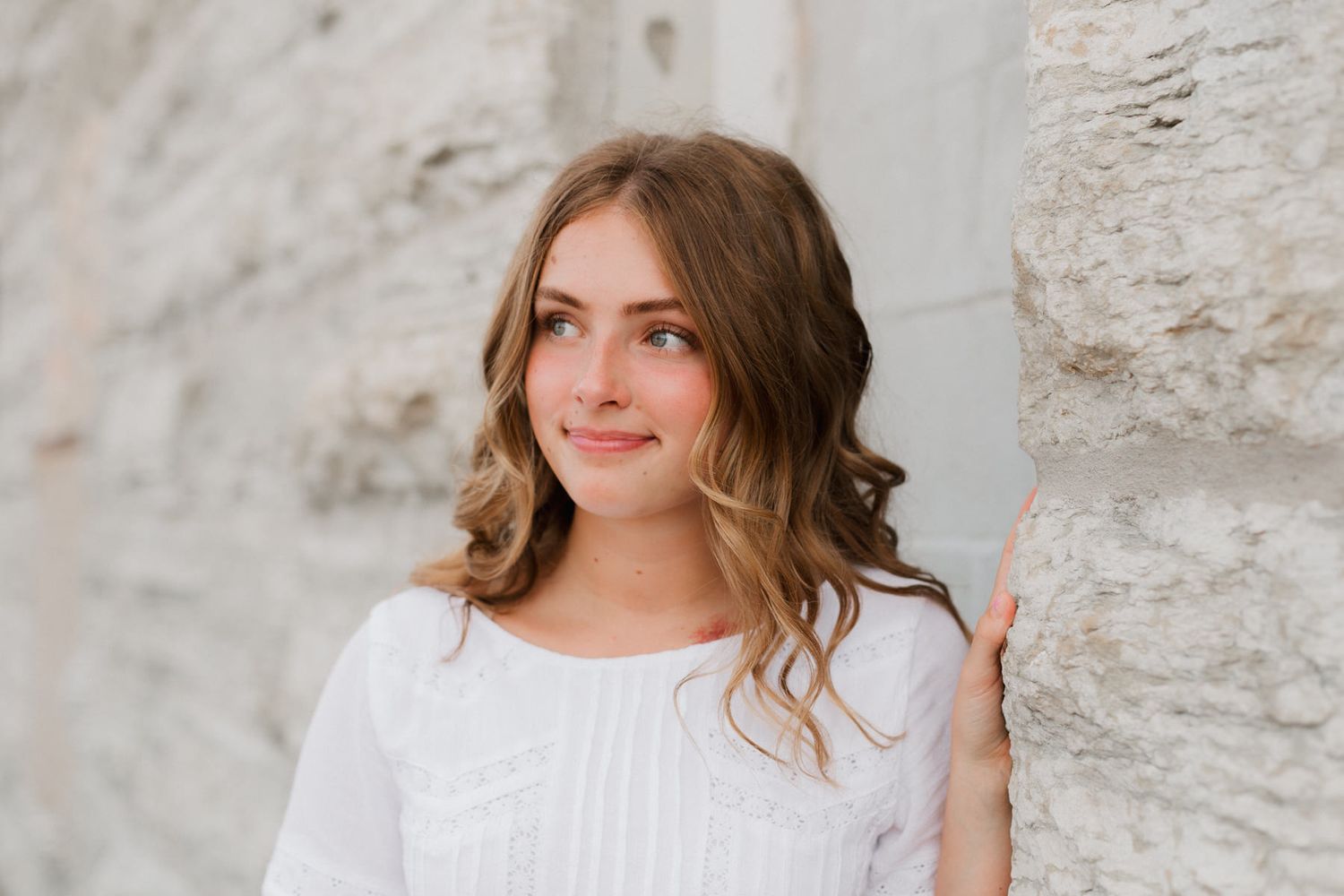 Teenage girl in white top leaning against stone wall, looking away softly.