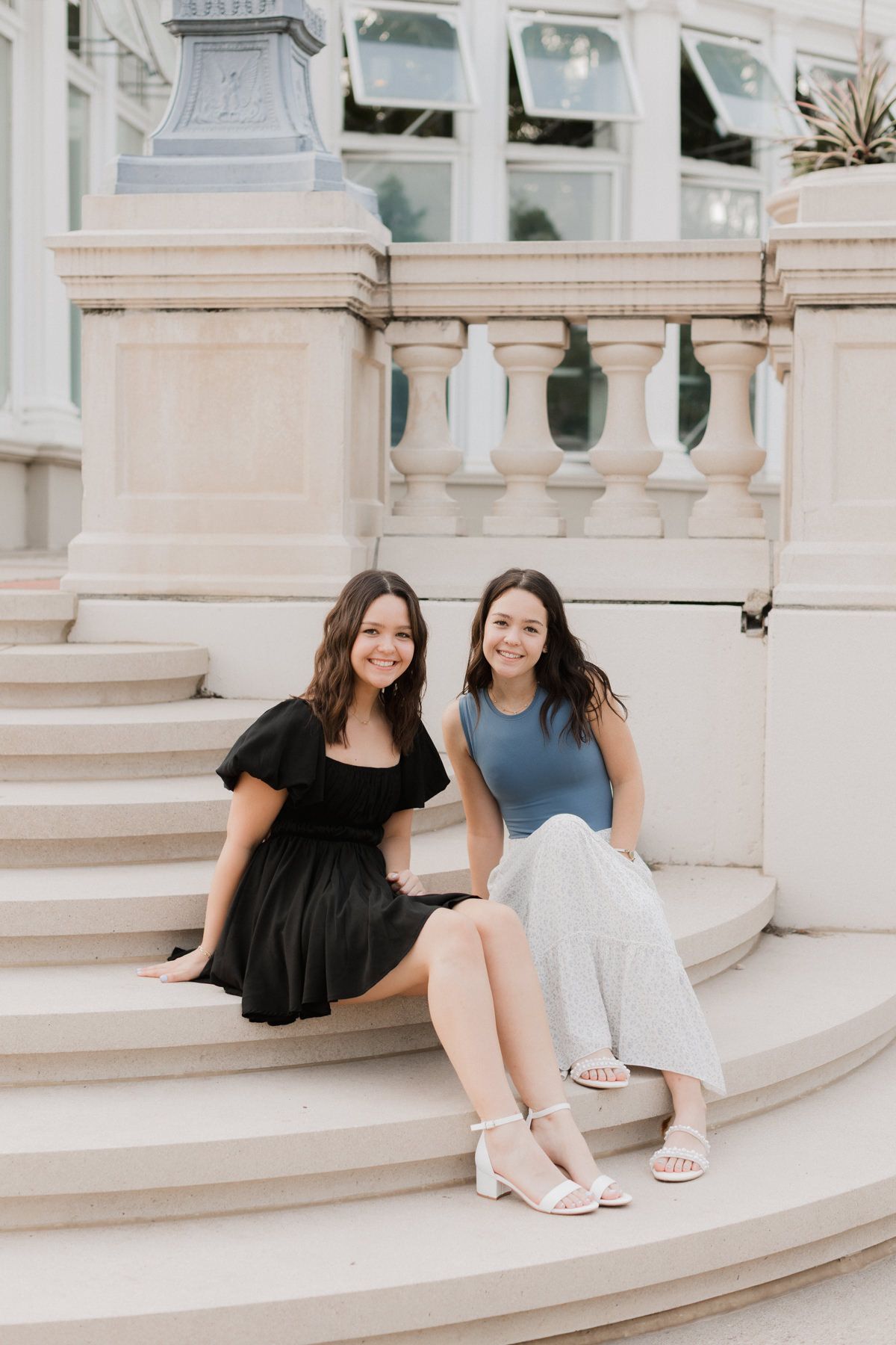 Two young women smiling and sitting on elegant stone steps outside a classical building with balustrades.