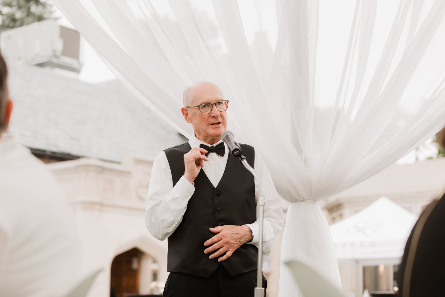 Bald man in black vest and bow tie adjusting his collar at an outdoor wedding ceremony with white draping.