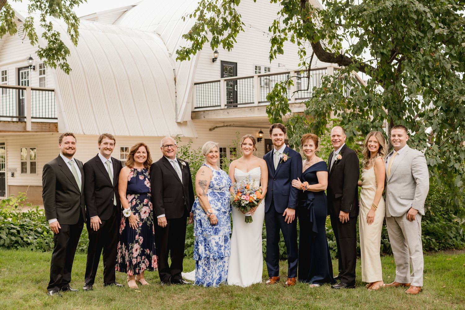 Wedding party group photo outdoors with bride and groom surrounded by family smiling in front of a white building.