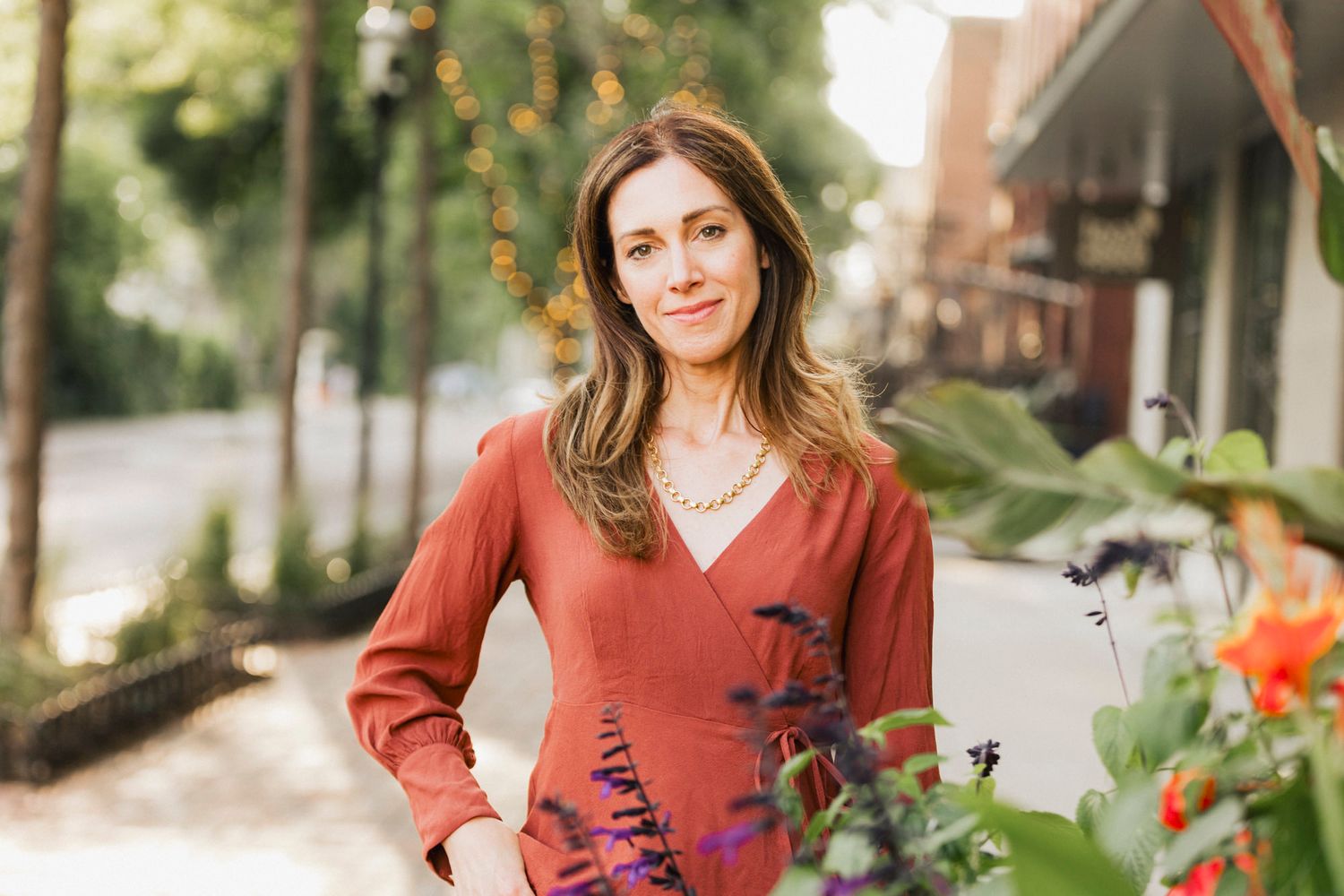 Outdoor portrait of person in rust-colored sweater standing near flowering plants.