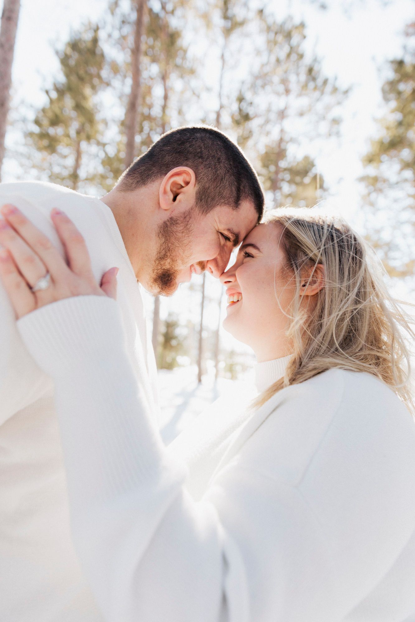 Intimate winter portrait of a couple in white sweaters touching foreheads in sunny forest setting.