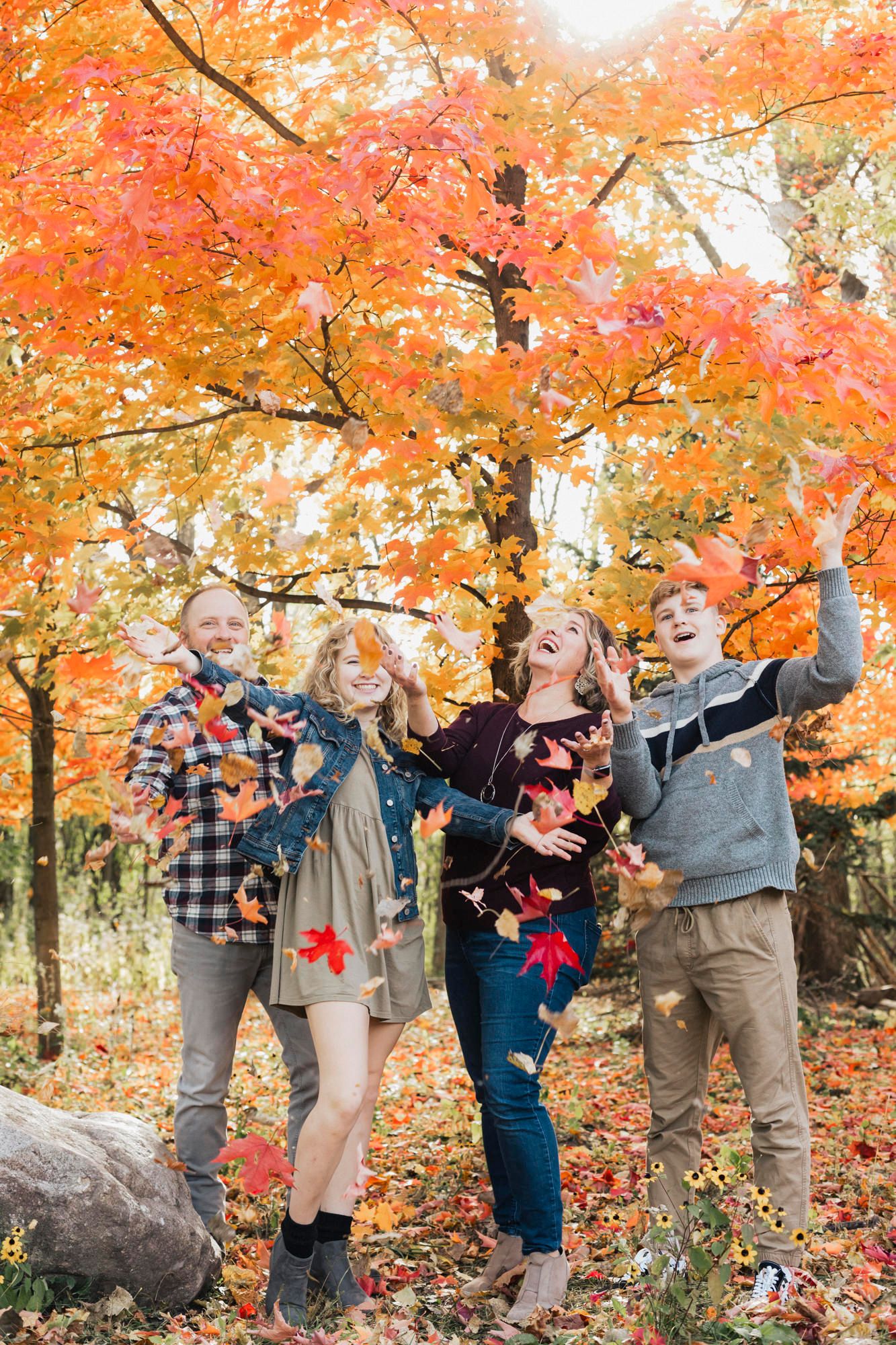 A group playing with falling autumn leaves under a vibrant orange maple tree.