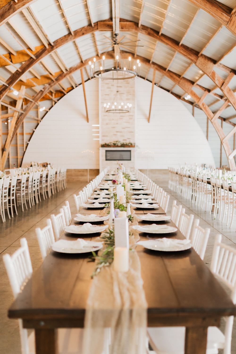 Long wooden banquet tables set for a wedding reception in a rustic barn venue with vaulted ceilings and natural light.