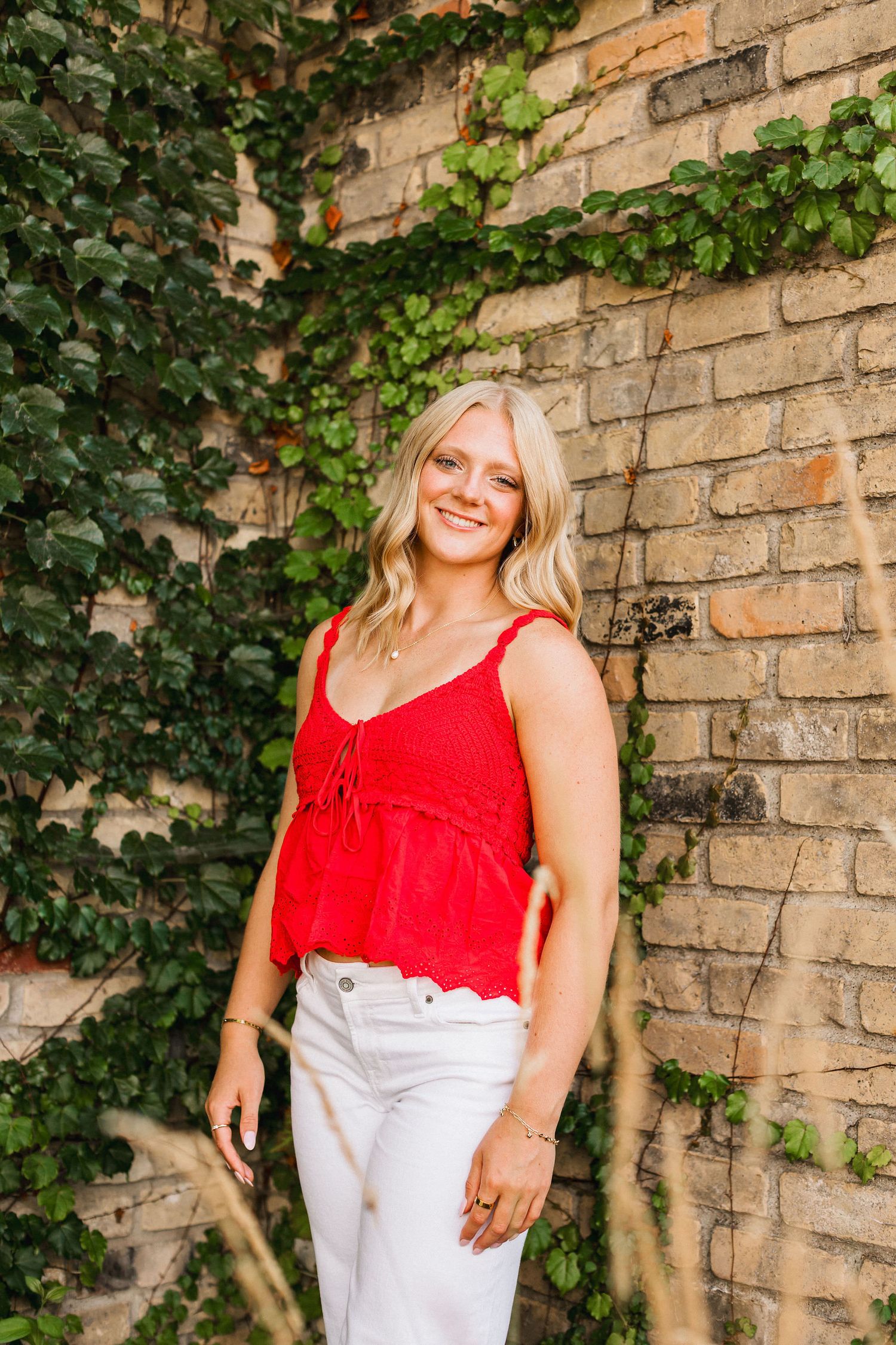 A summer photoshoot against an ivy-covered brick wall featuring a red tank top and white pants outfit.
