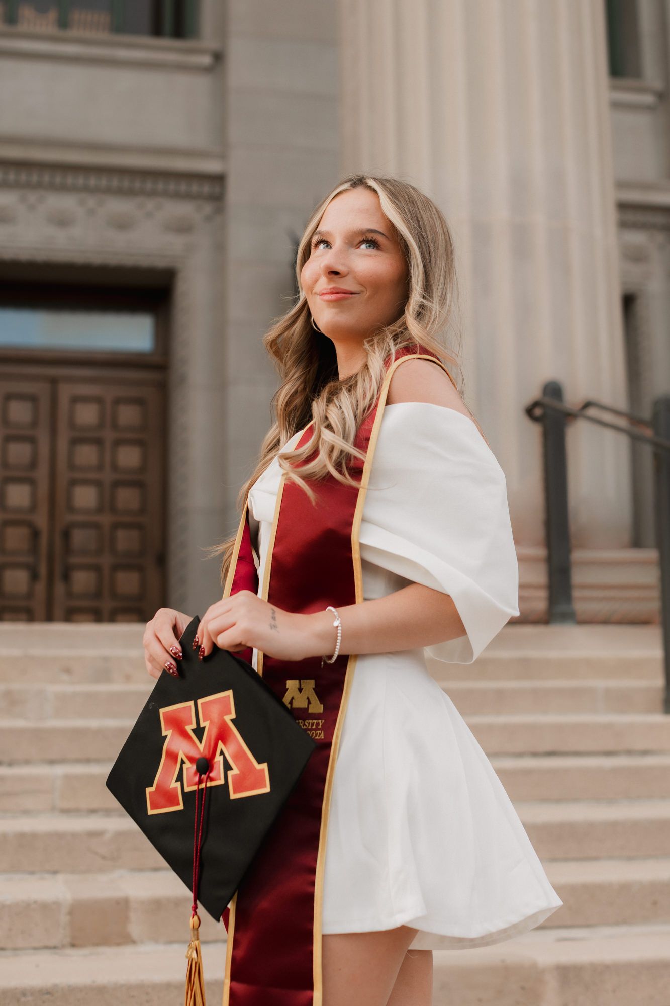 Blonde woman in white dress holding University of Minnesota graduation cap on university steps.