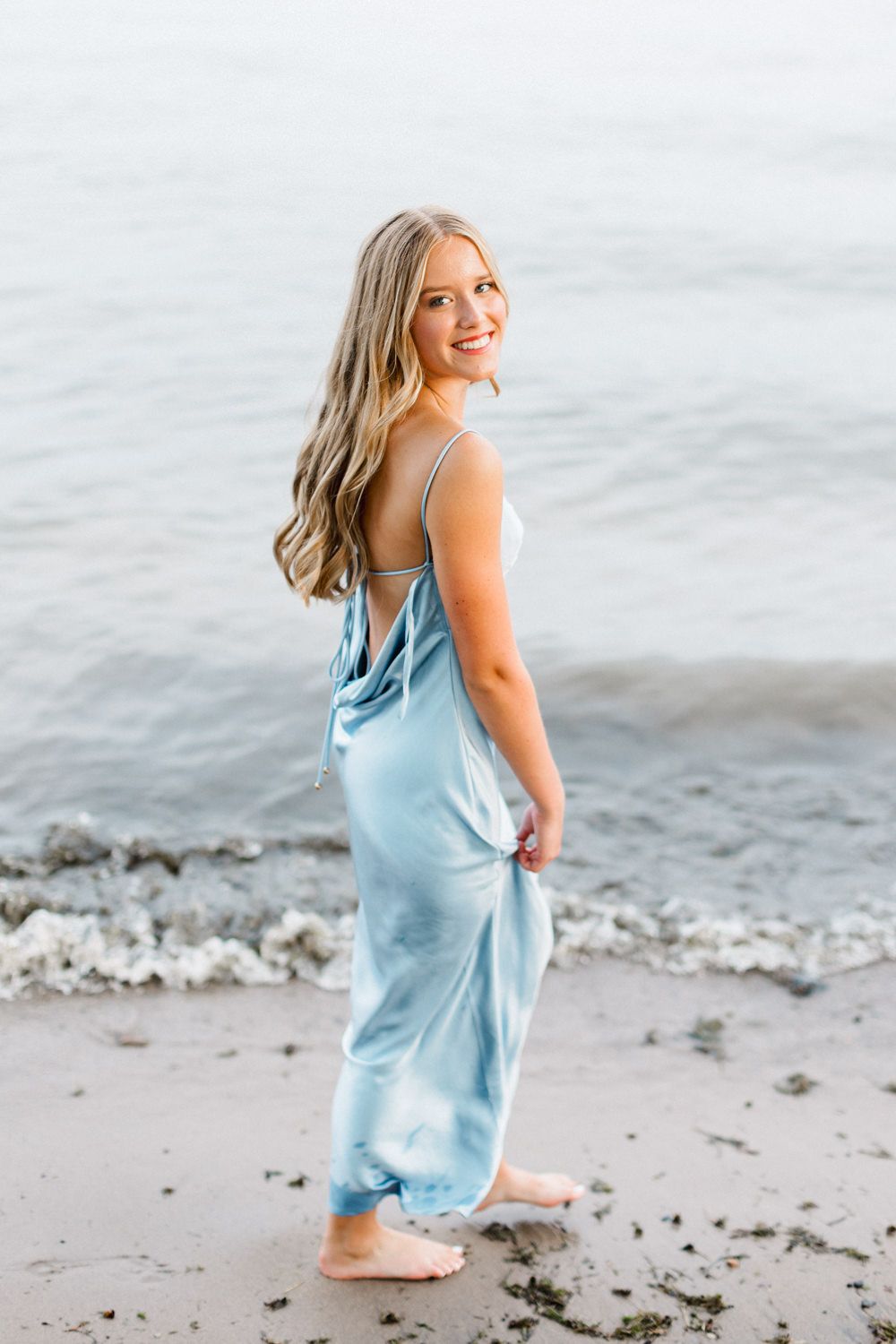 A person in a pastel blue dress poses barefoot on the beach with waves in the background.
