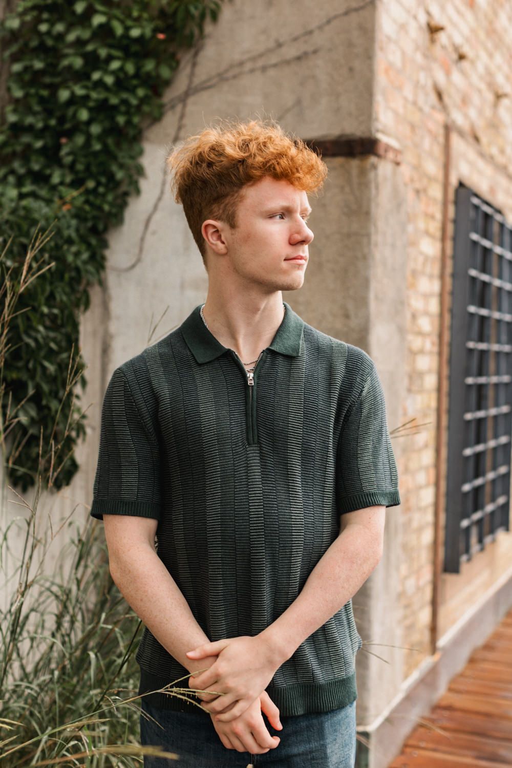 A person in a dark green polo shirt poses against a brick wall with climbing vines.