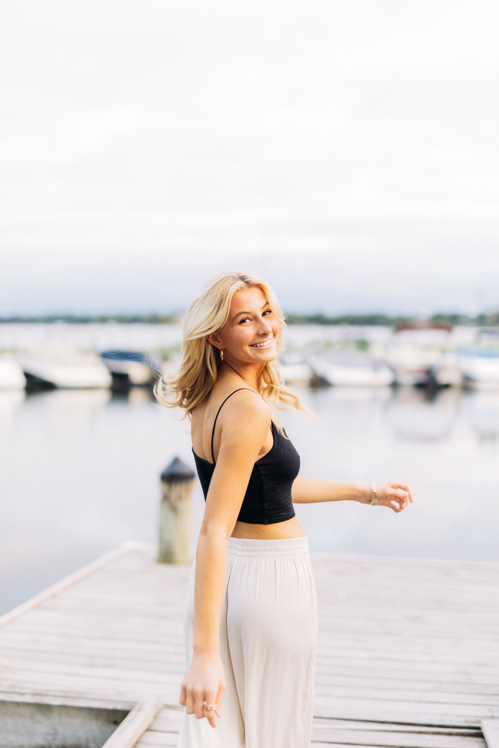 Woman in a black top and white skirt dances playfully on a wooden dock by the water on a sunny day.