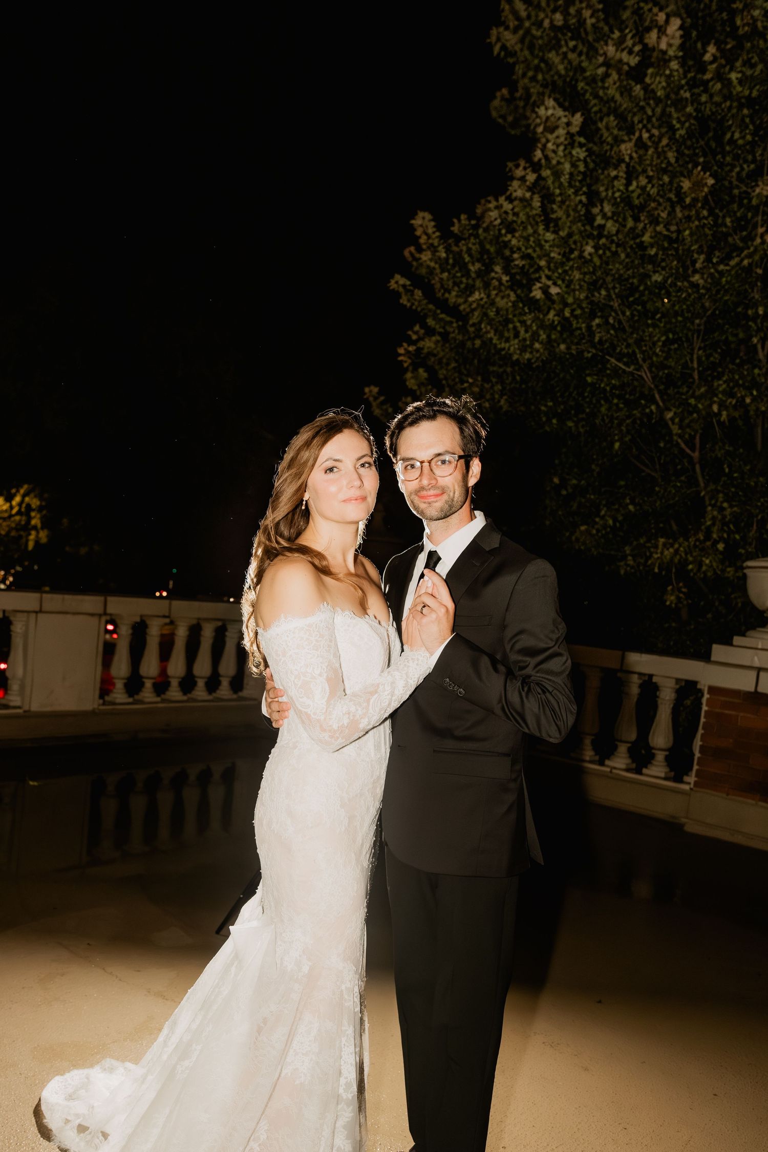 A bride in a lace mermaid gown and groom in a black tuxedo pose romantically outdoors at night.