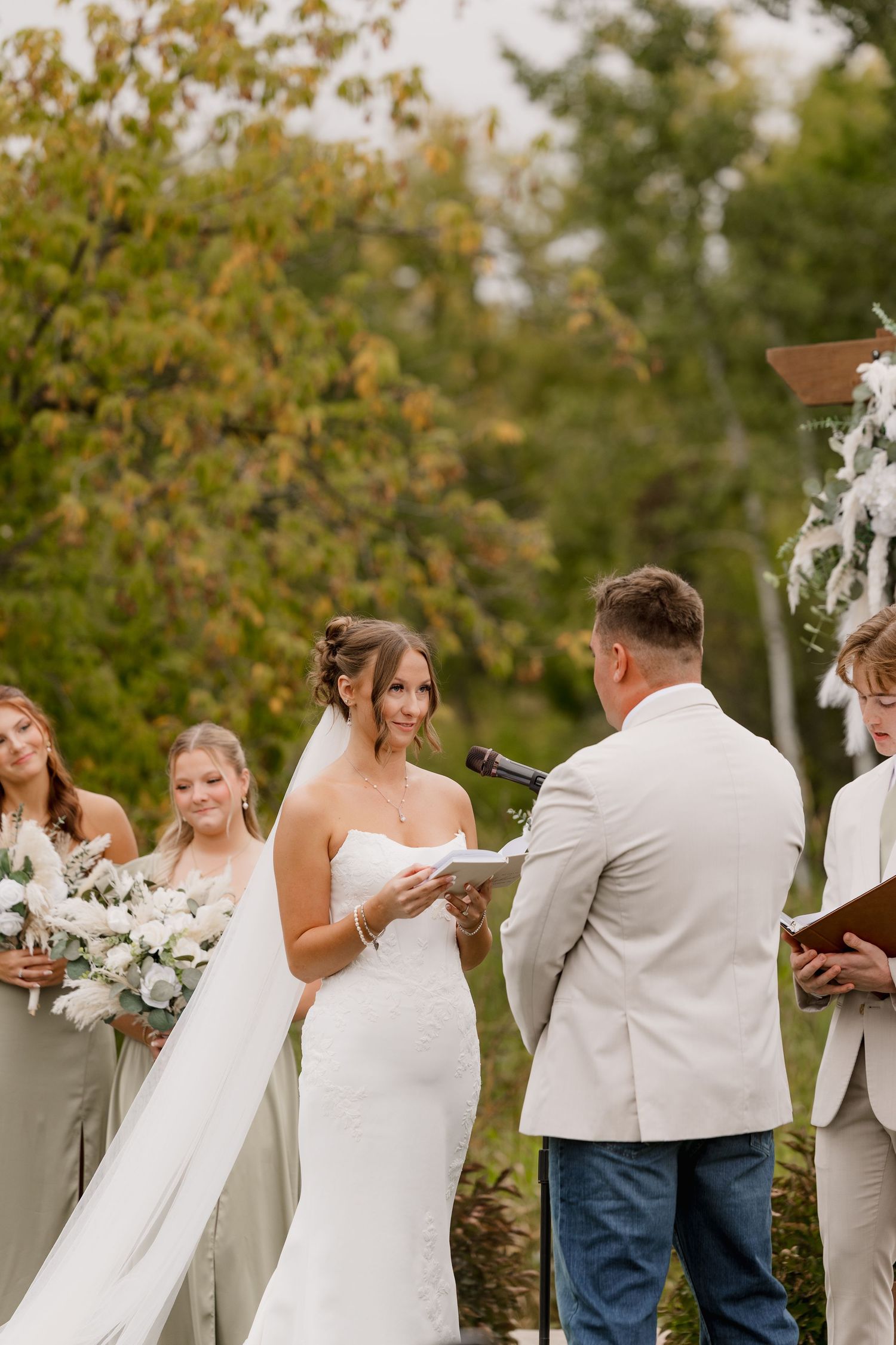 Bride in white gown reads vows to groom at outdoor wedding ceremony with bridesmaids behind her.