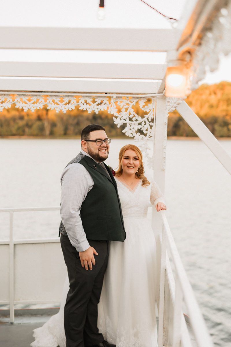 A romantic wedding couple poses together on a white boat deck with string lights and water views.