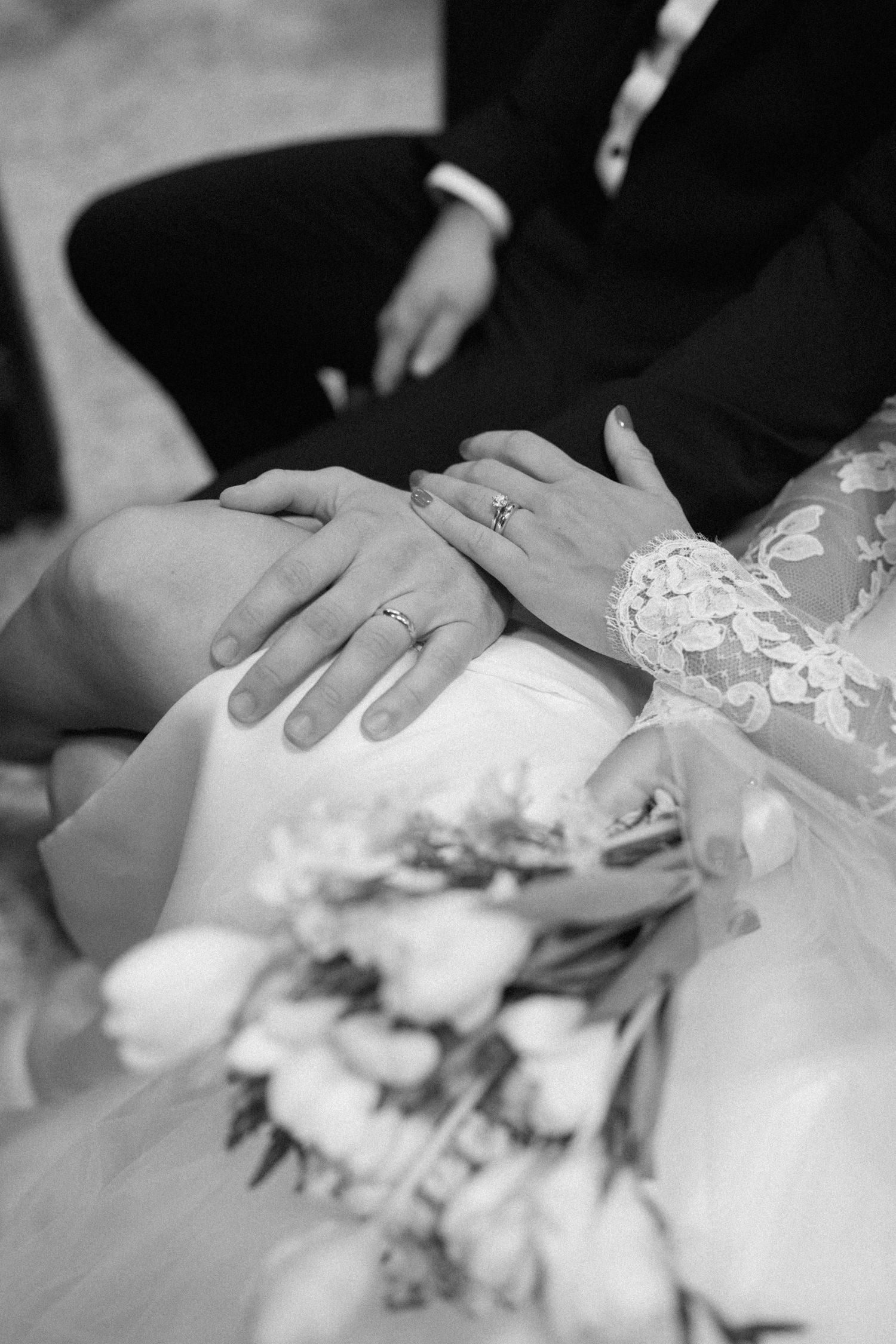 Close-up black and white photo of wedding rings and hands resting on a lace dress with flowers.