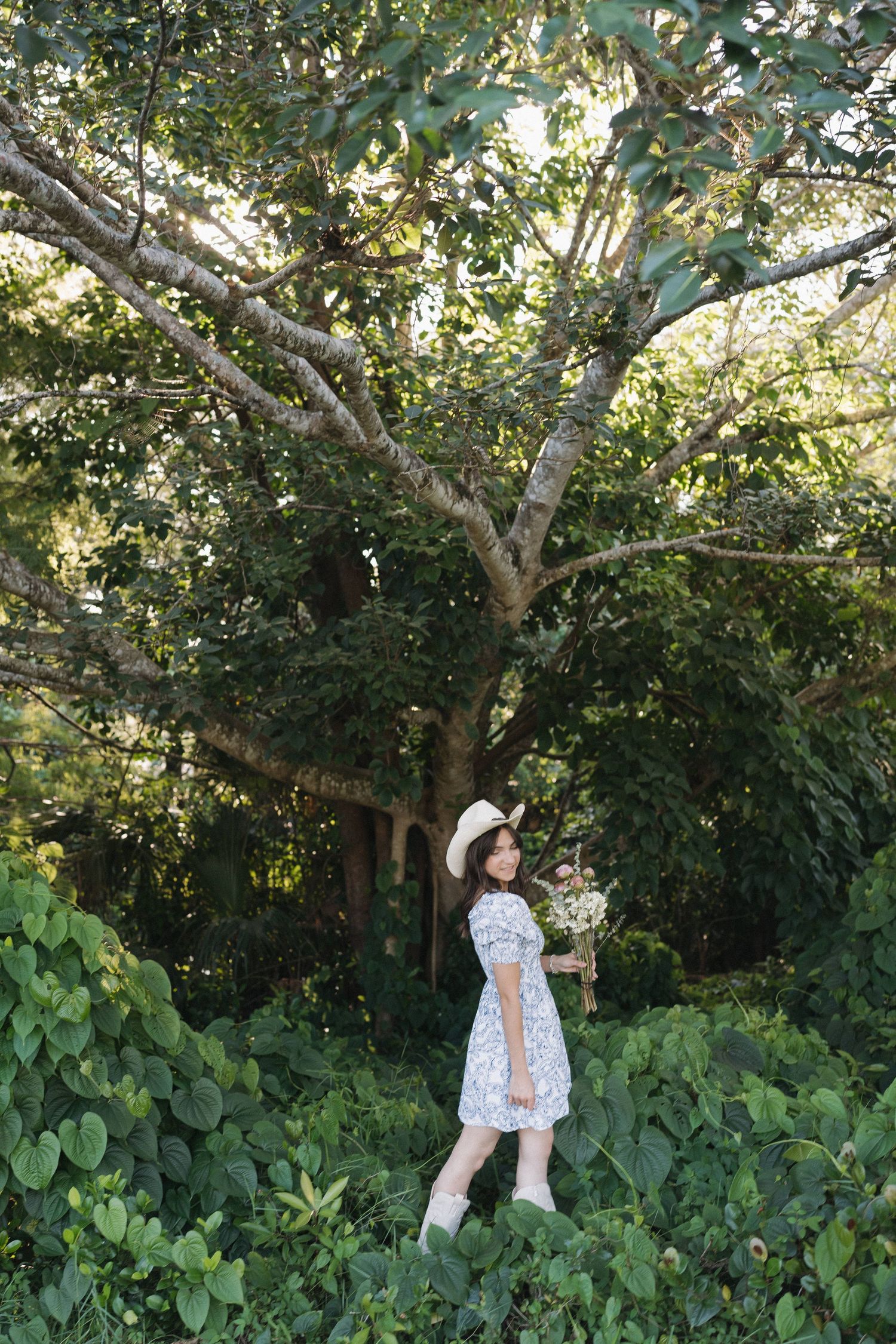 White sundress and hat silhouette poses near lush green trees in a summer garden setting.