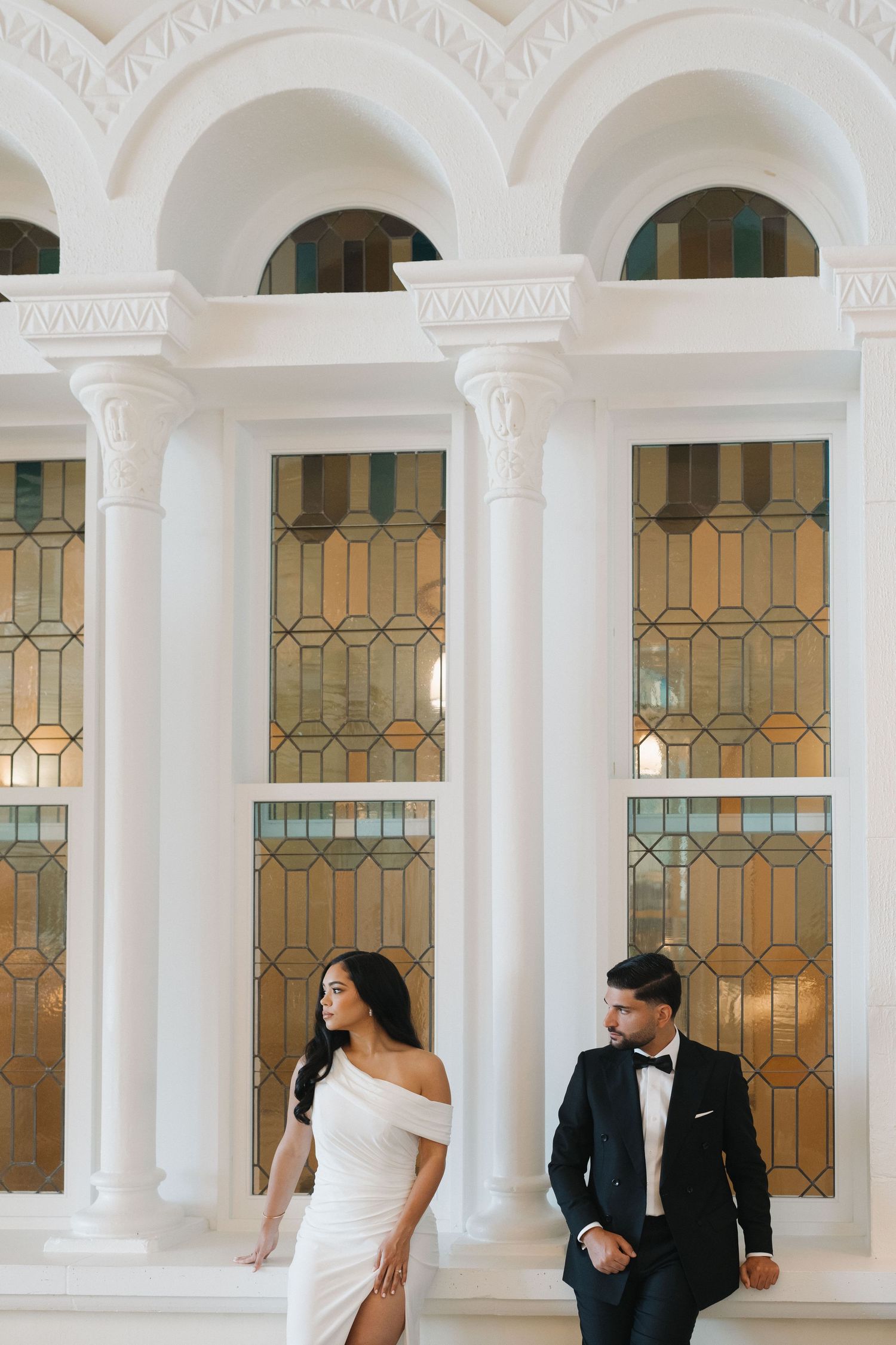 Elegant couple poses in formal attire against a backdrop of white columns and stained glass windows.
