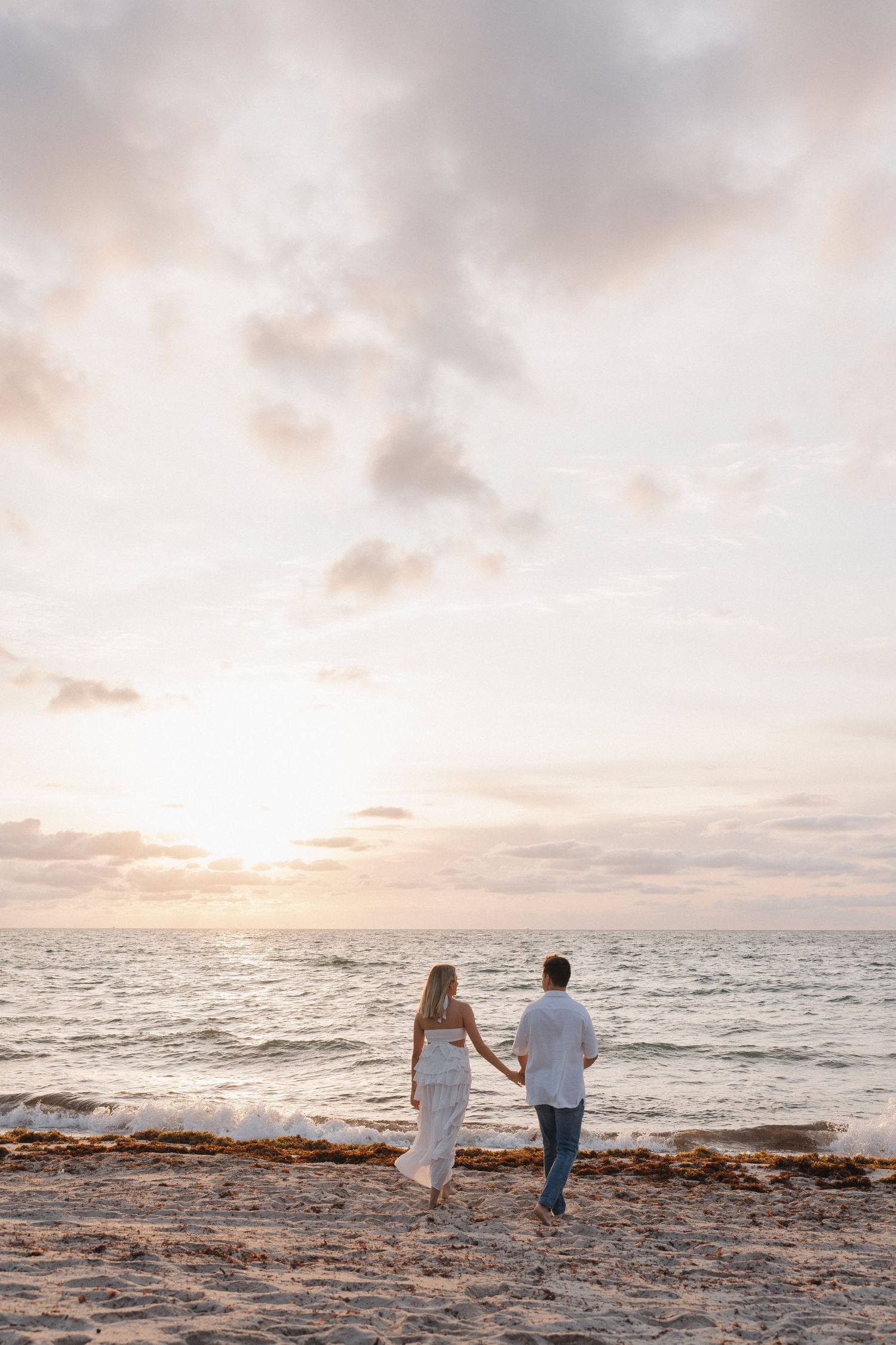 Couple walking hand in hand on a sandy beach towards the setting sun over the ocean.
