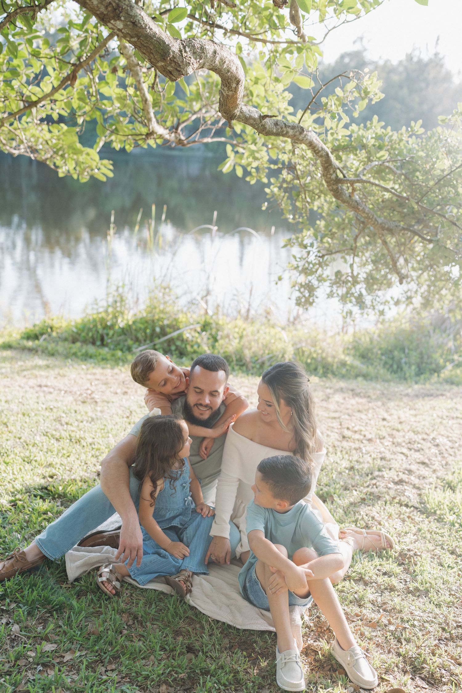 Family relaxing on blanket by lake with overhanging tree branches.