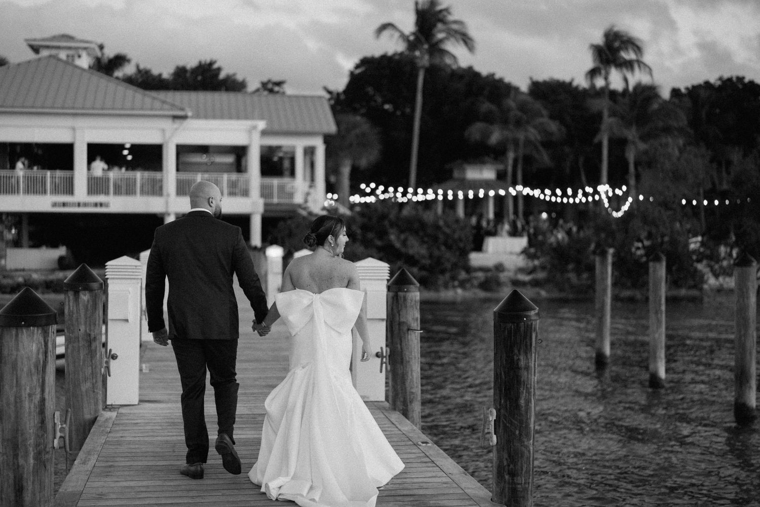 A black and white photo of newlyweds walking along a waterfront dock at dusk.