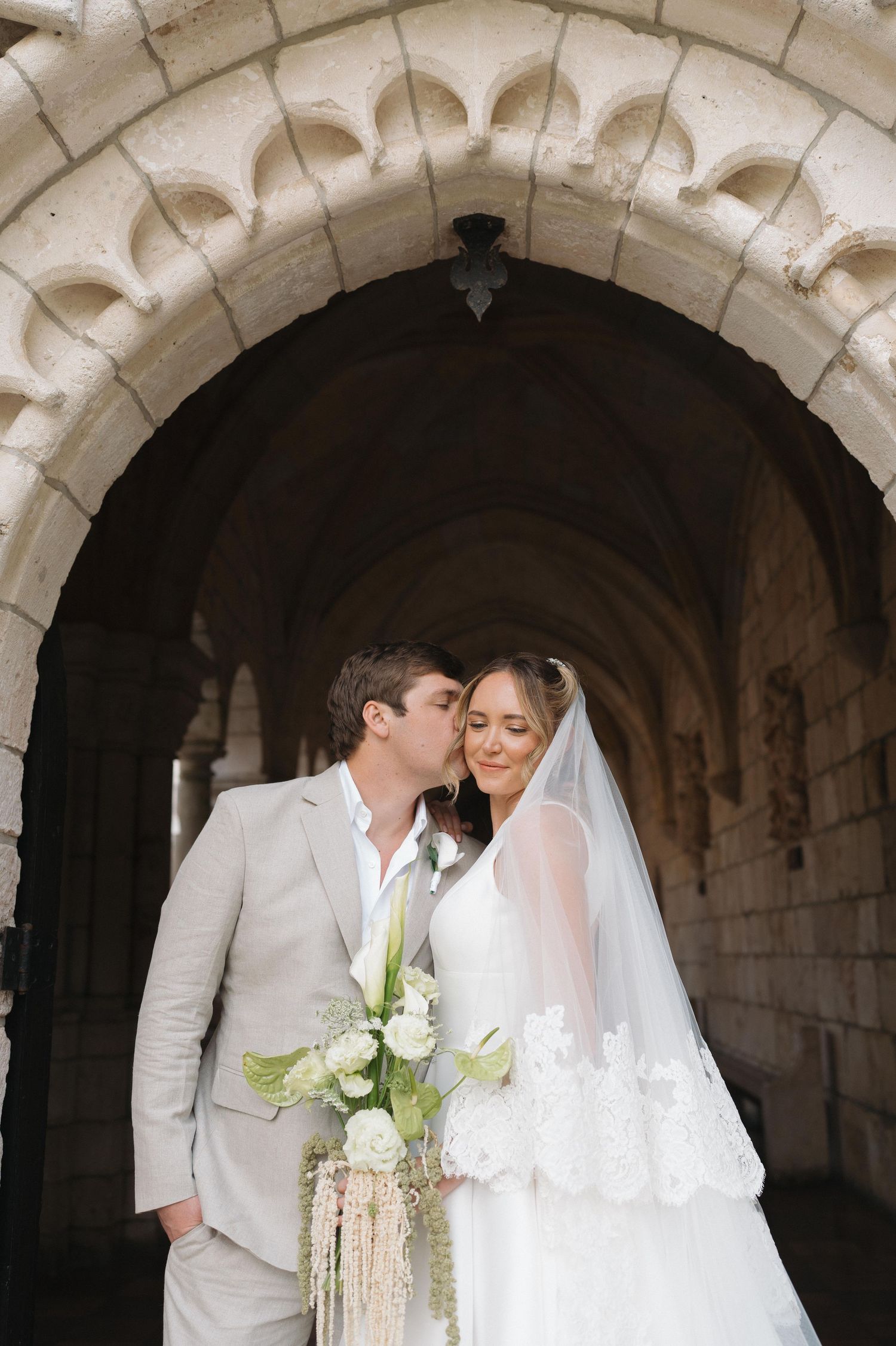 Romantic wedding portrait under a stone archway with white flowers and elegant attire.
