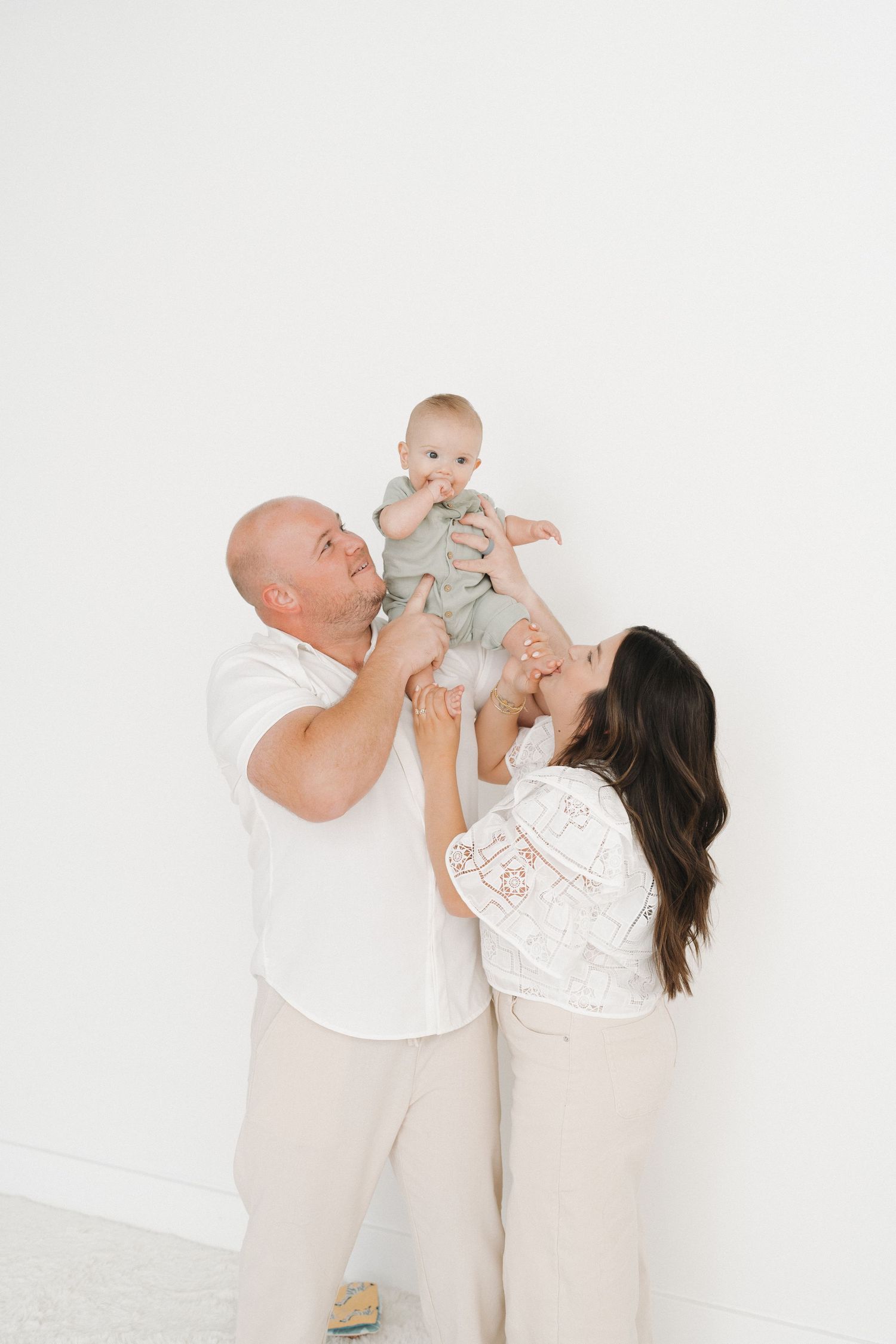 Family portrait with parents lifting baby in air against white wall.