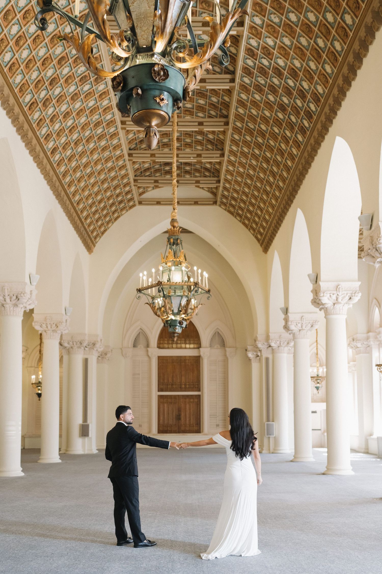 Couple holds hands while standing apart in an ornate hallway with arched ceilings and chandeliers.