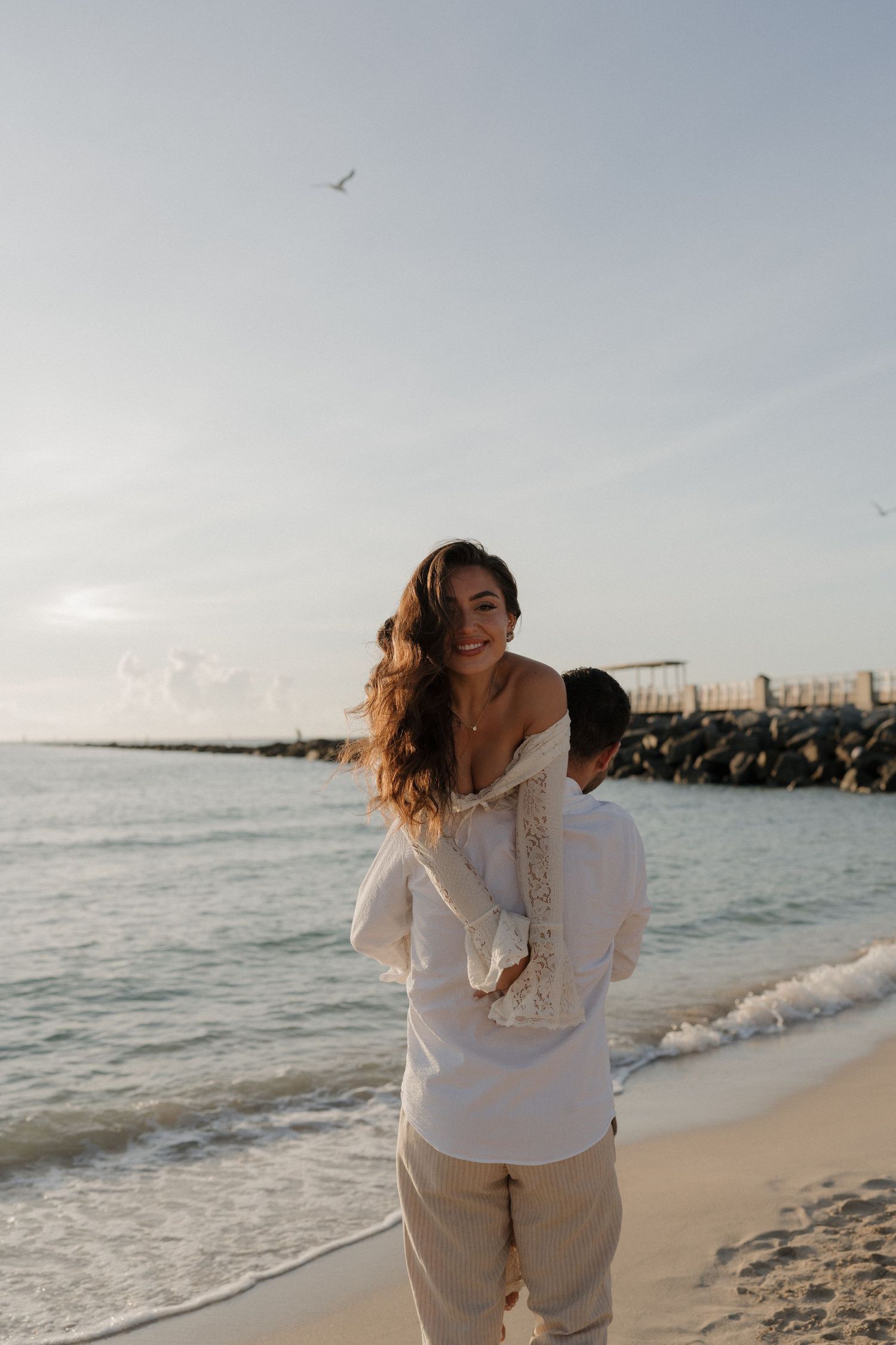 A playful moment captured as a couple enjoys a piggyback ride on the beach with seabirds flying overhead.