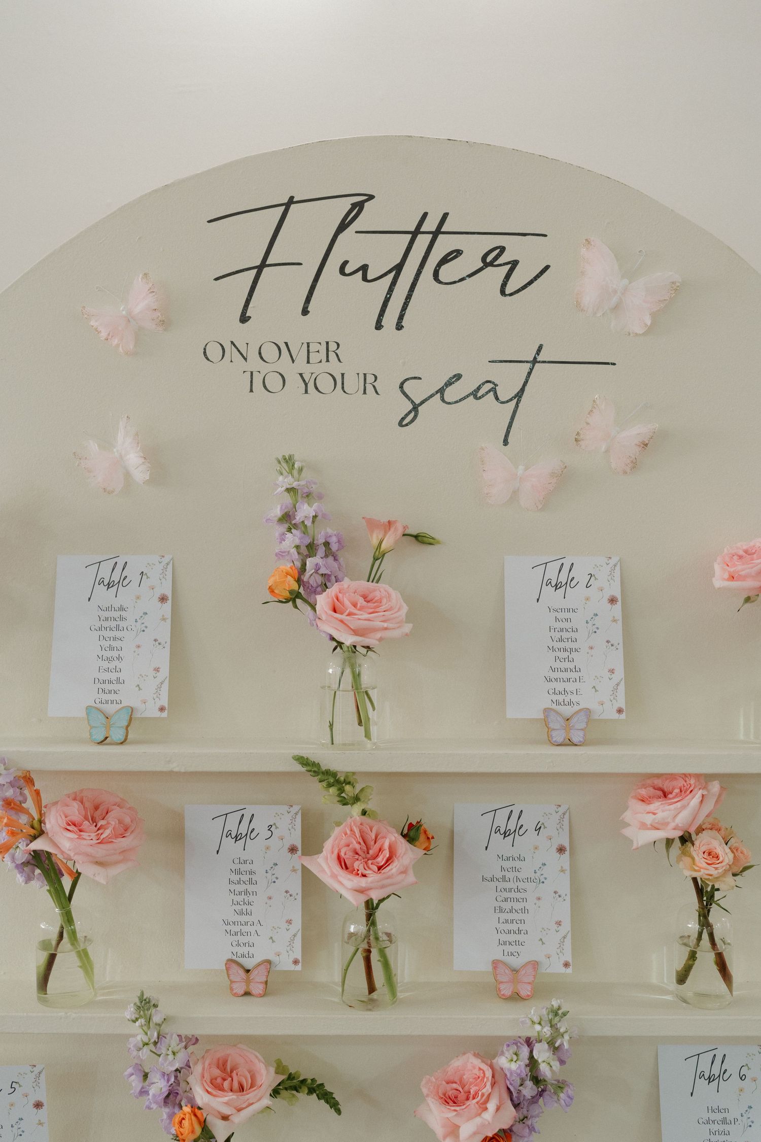 Wedding seating chart display with pink roses and butterflies under 'Flutter to your seat' text.