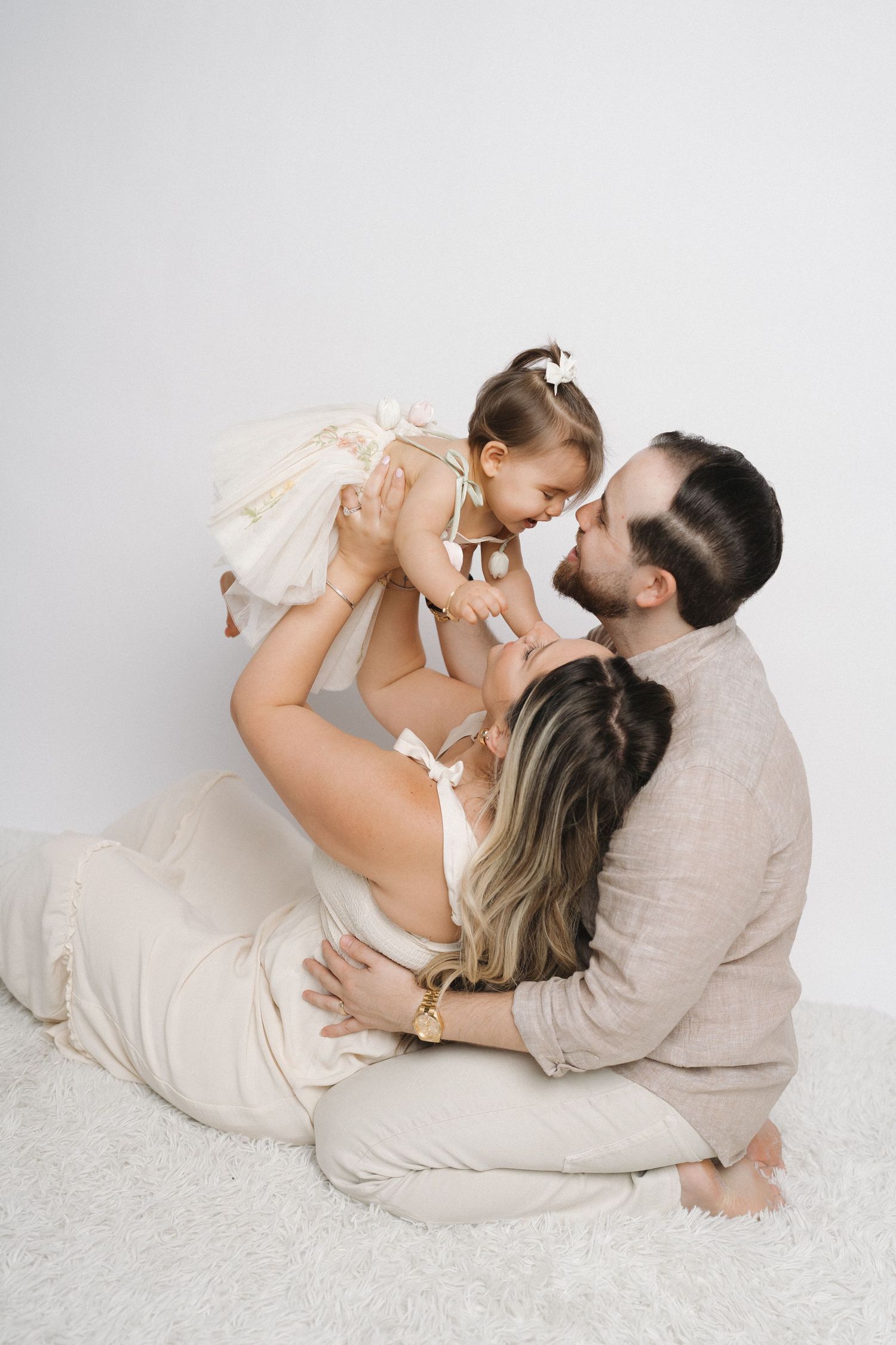 Family in neutral colors shares tender moment while laying on white carpet.