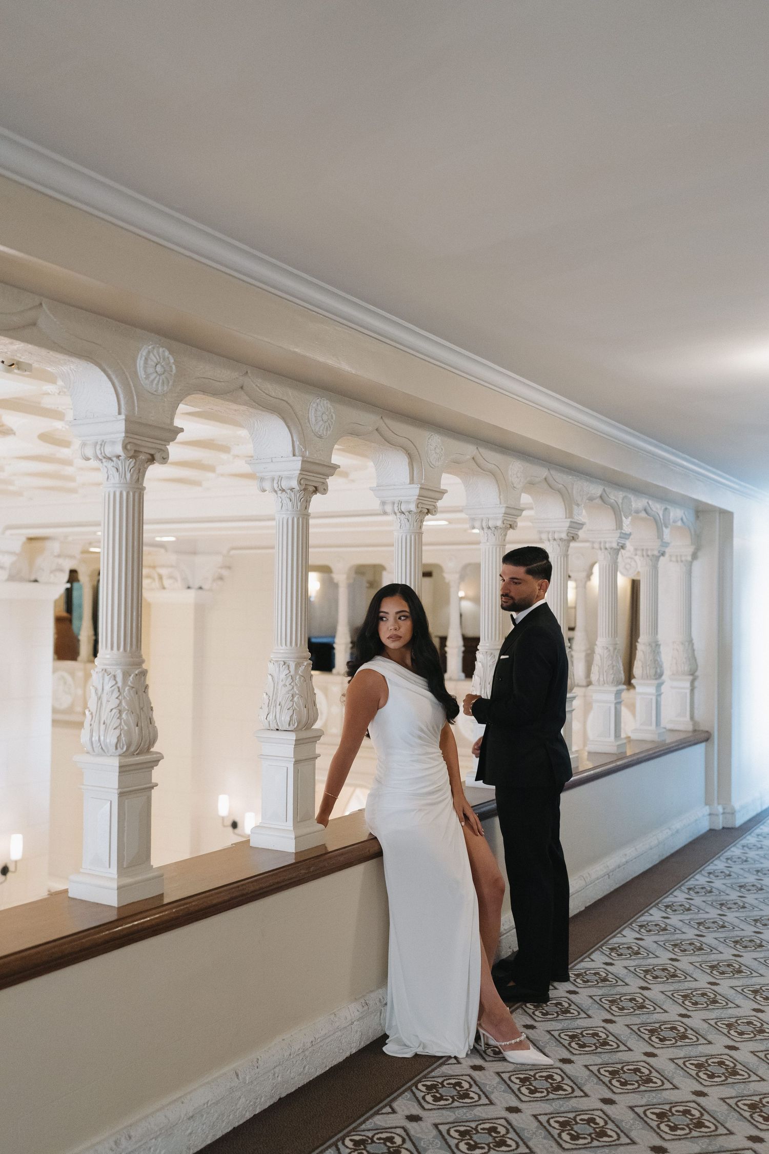 Elegant couple posing in ornate white hallway with classical columns and patterned floor.