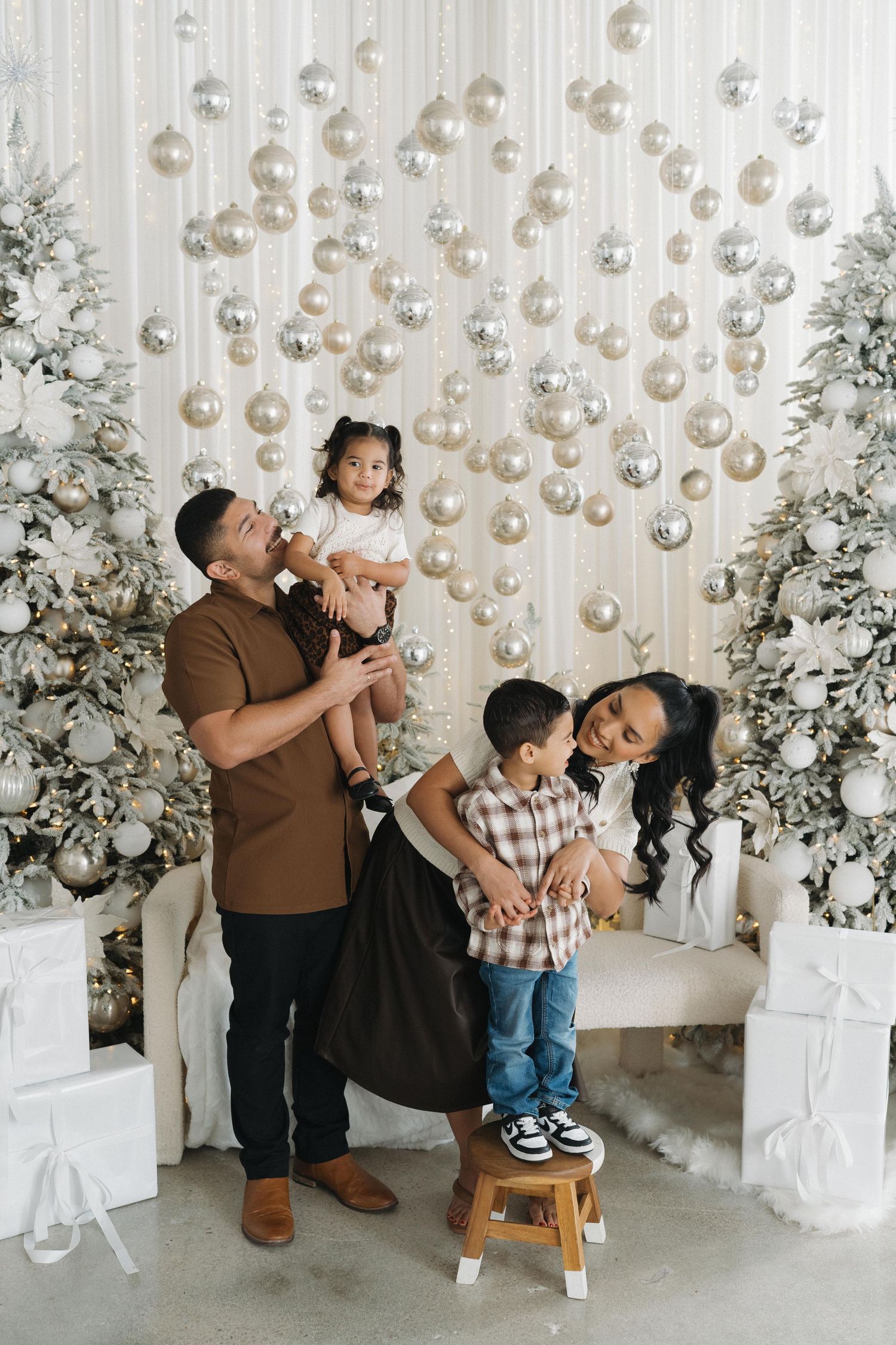 Family of four posing in festive Christmas studio with ornament backdrop, flocked trees, and white gifts.