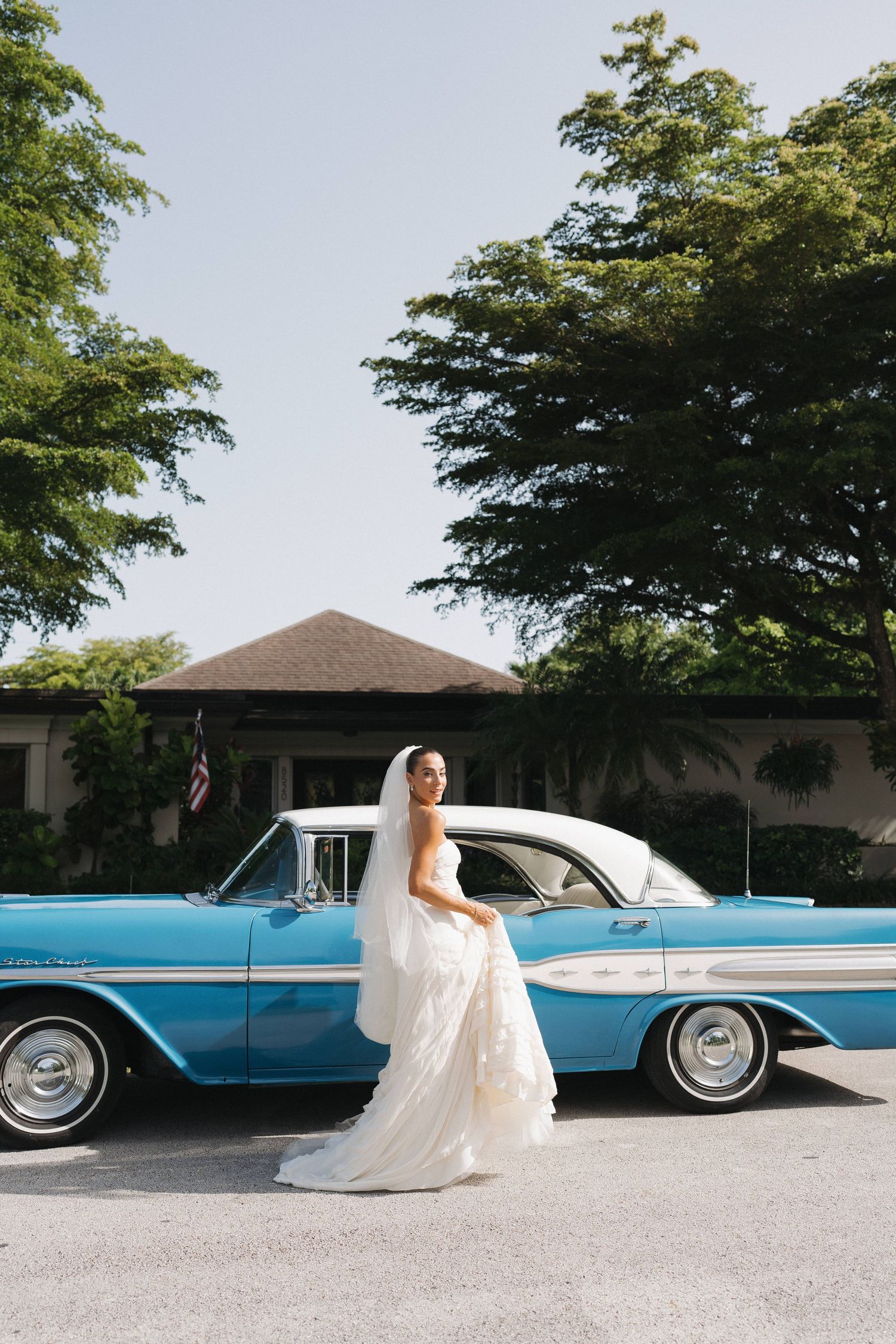 Bride in white dress poses beside vintage blue car on sunny day.