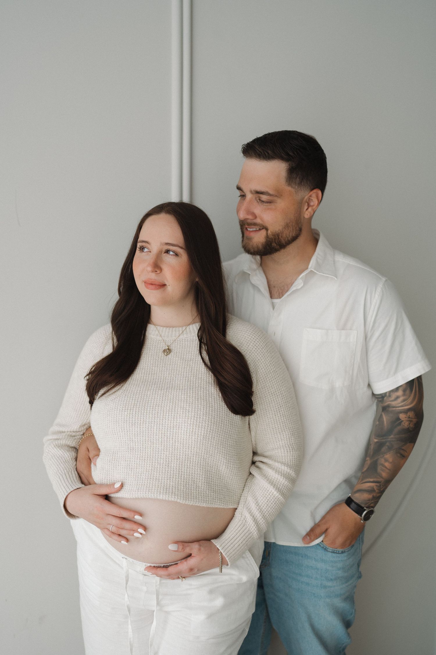 Couple in casual white outfits sharing intimate maternity moment against minimalist wall.