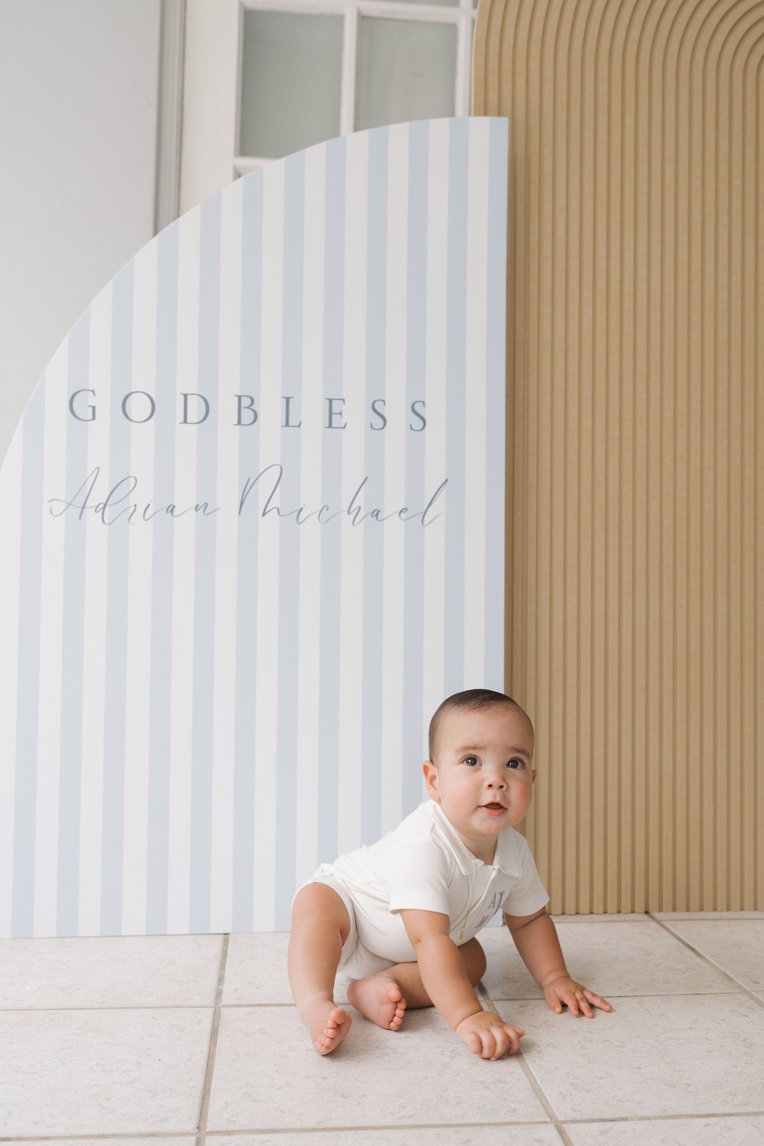 Baby in white onesie crawling across floor with soft blue striped wall backdrop and 'God Bless' text.