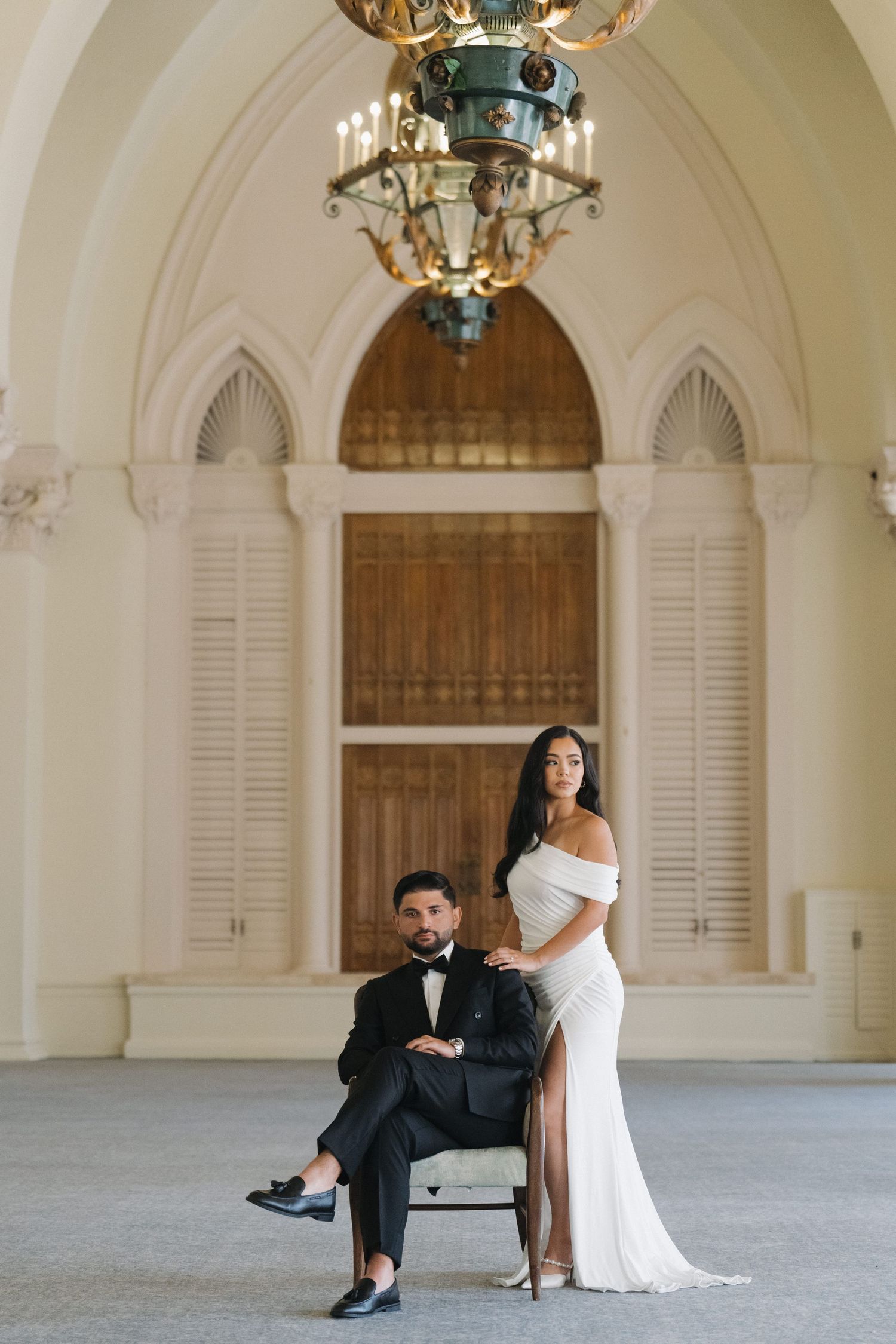 A couple shares an intimate moment in a grand room with arched windows and ornate chandeliers.