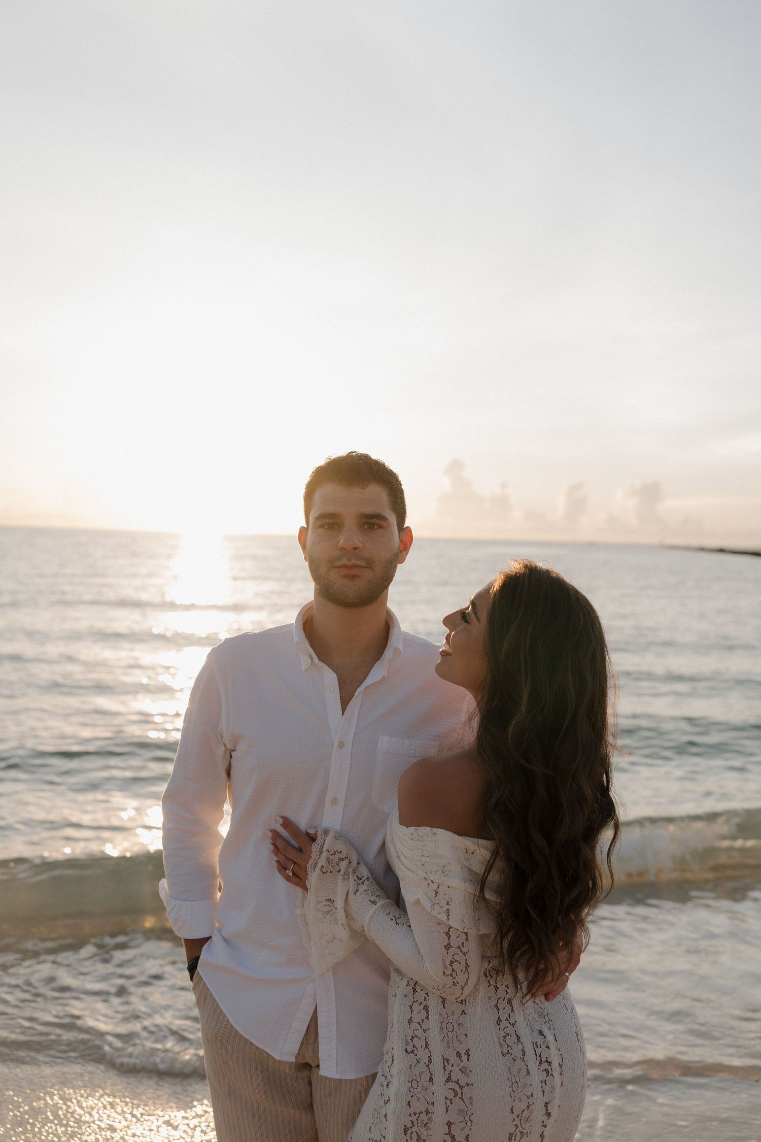 A romantic couple poses at sunset on a beach with the ocean and glowing sun in the background.