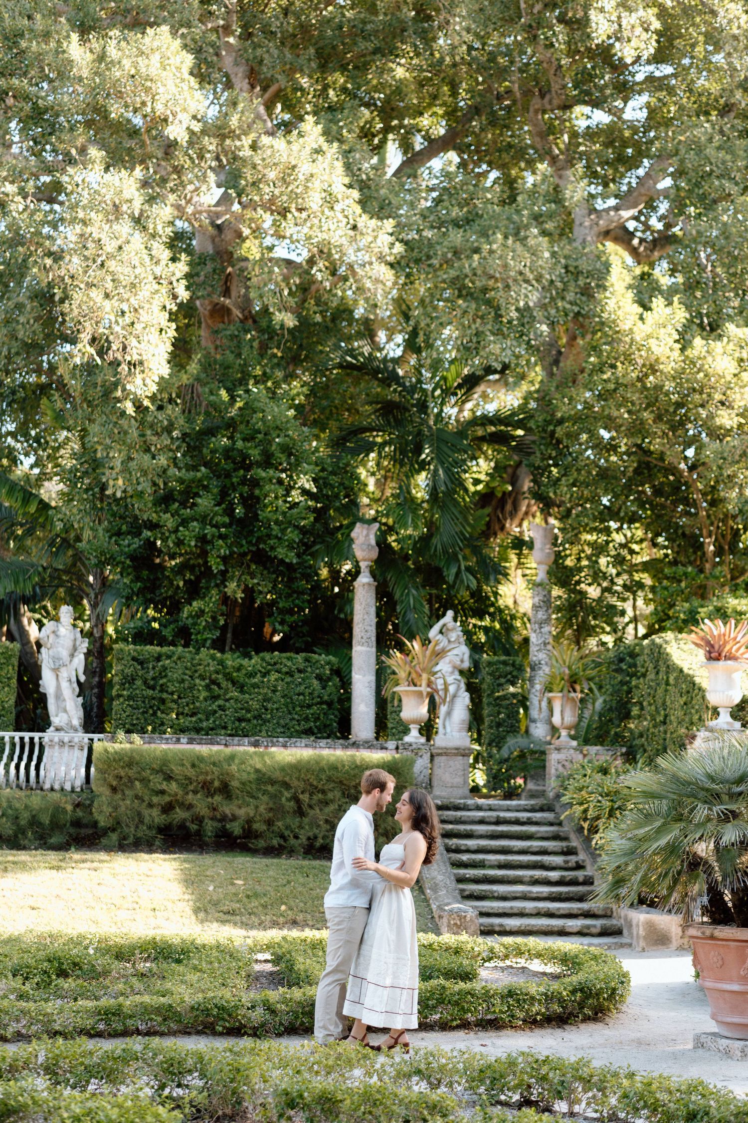 A couple embraces in a manicured garden with blooming trees and classical statuary.